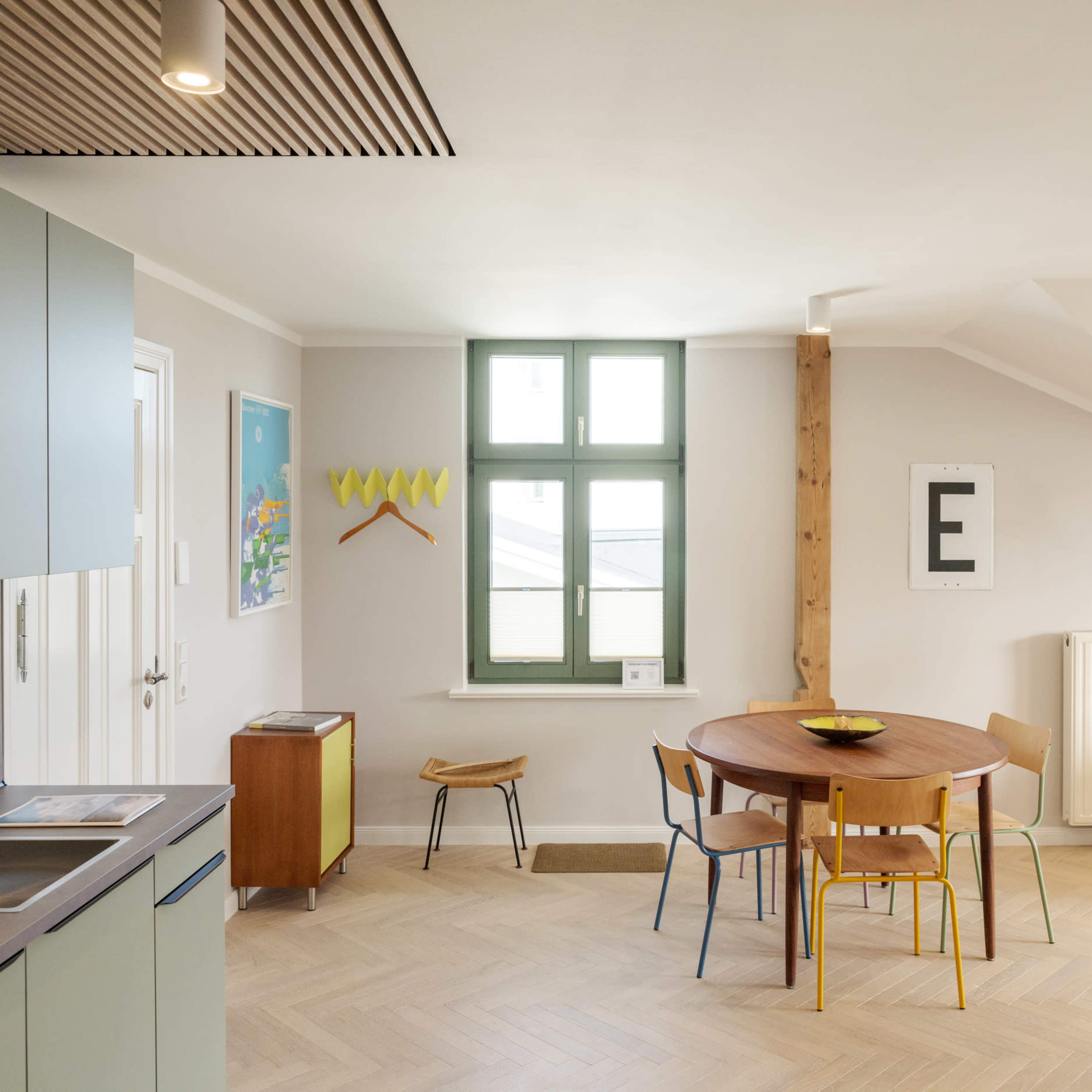 A modern living space features a round wooden table surrounded by colorful chairs, with a kitchen area in the background and a cozy sofa in the corner.