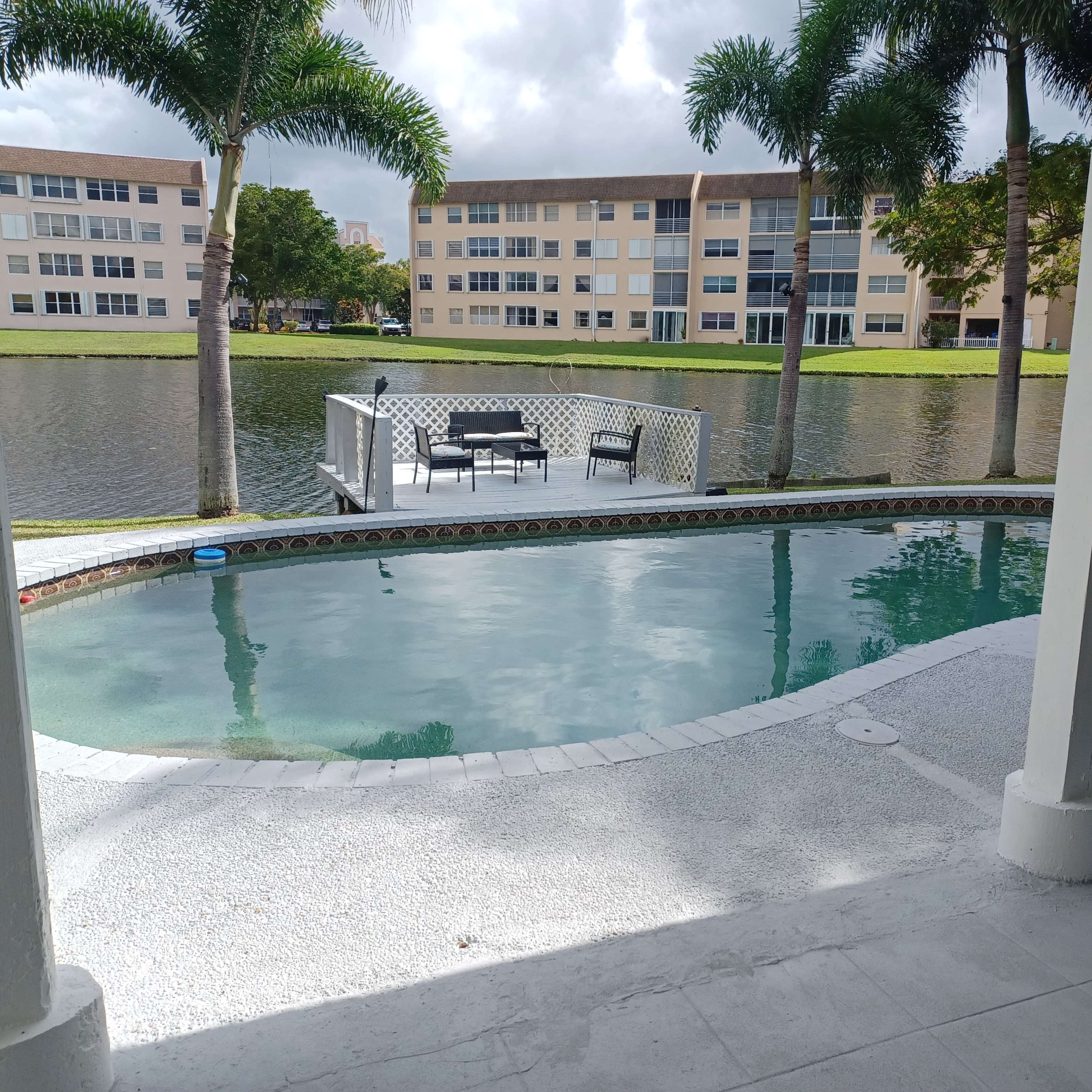 A residential pool area with a view of a lake and nearby buildings, featuring lounge chairs and palm trees.