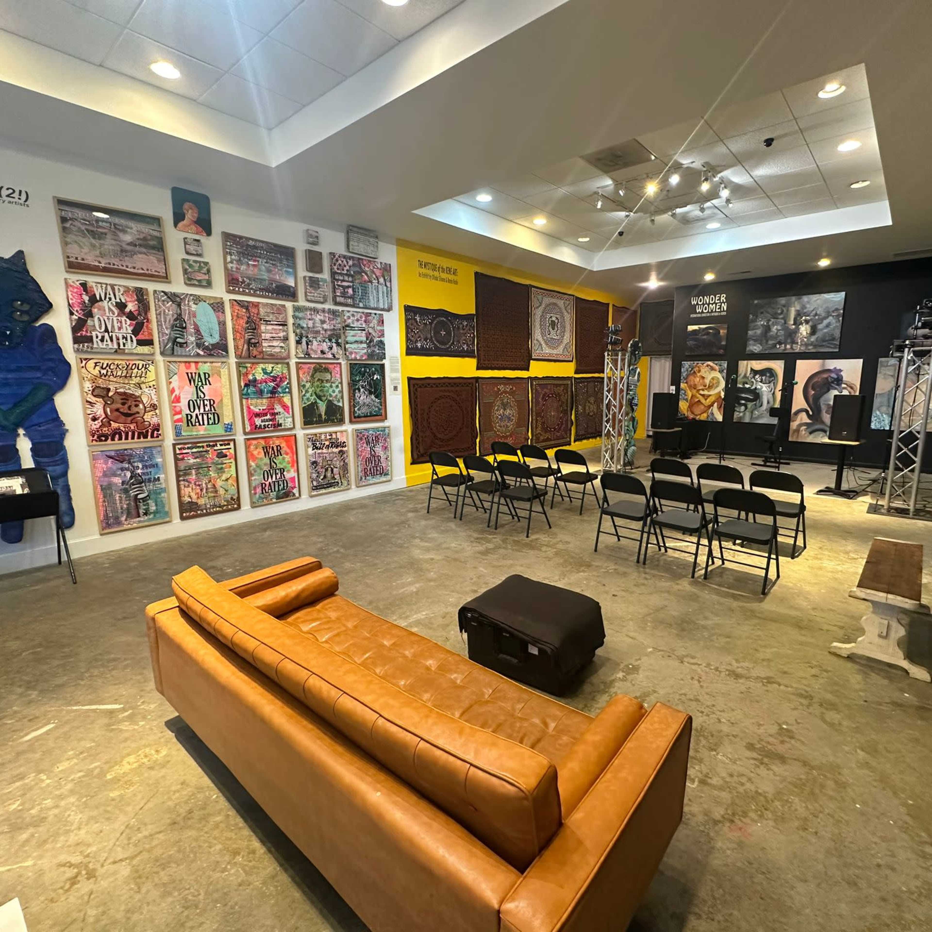 An art gallery space with a brown leather couch, several colorful art pieces on the walls, and rows of black chairs arranged for an event.