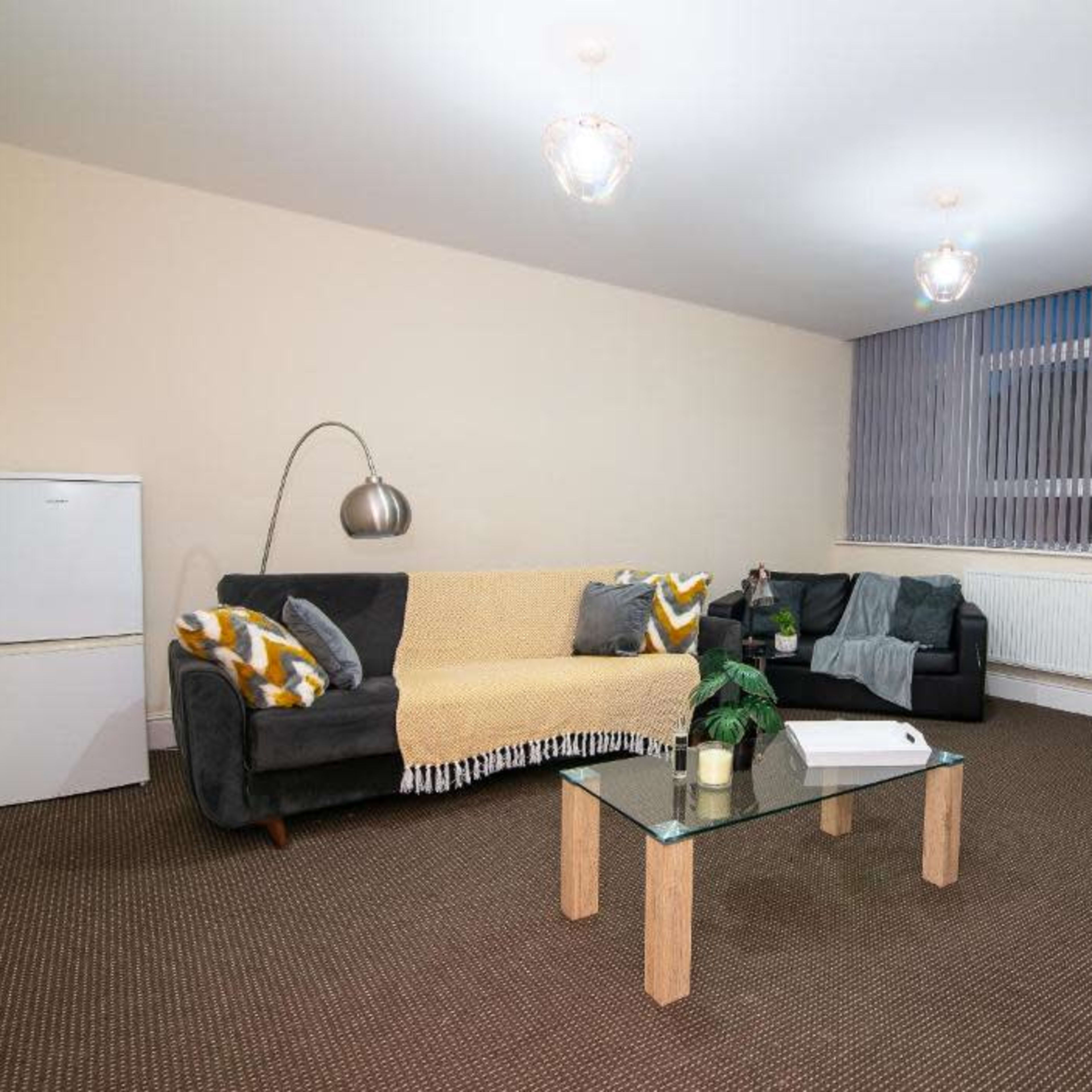 A minimalist living room features a gray couch with a yellow throw, a glass coffee table, a white refrigerator, and large windows with vertical blinds.