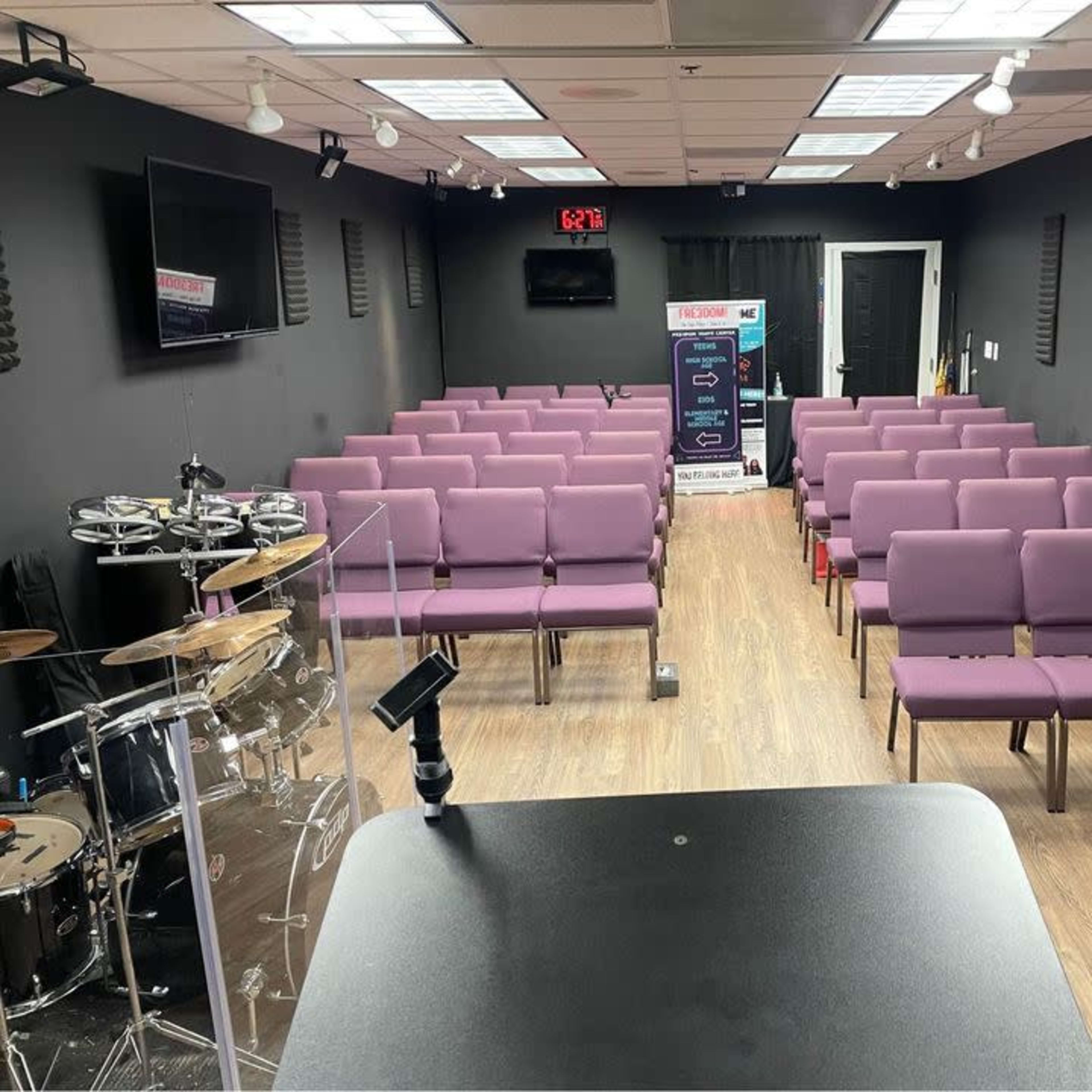 A spacious room arranged with purple seating facing a central stage area, with music equipment on one side and a black backdrop.