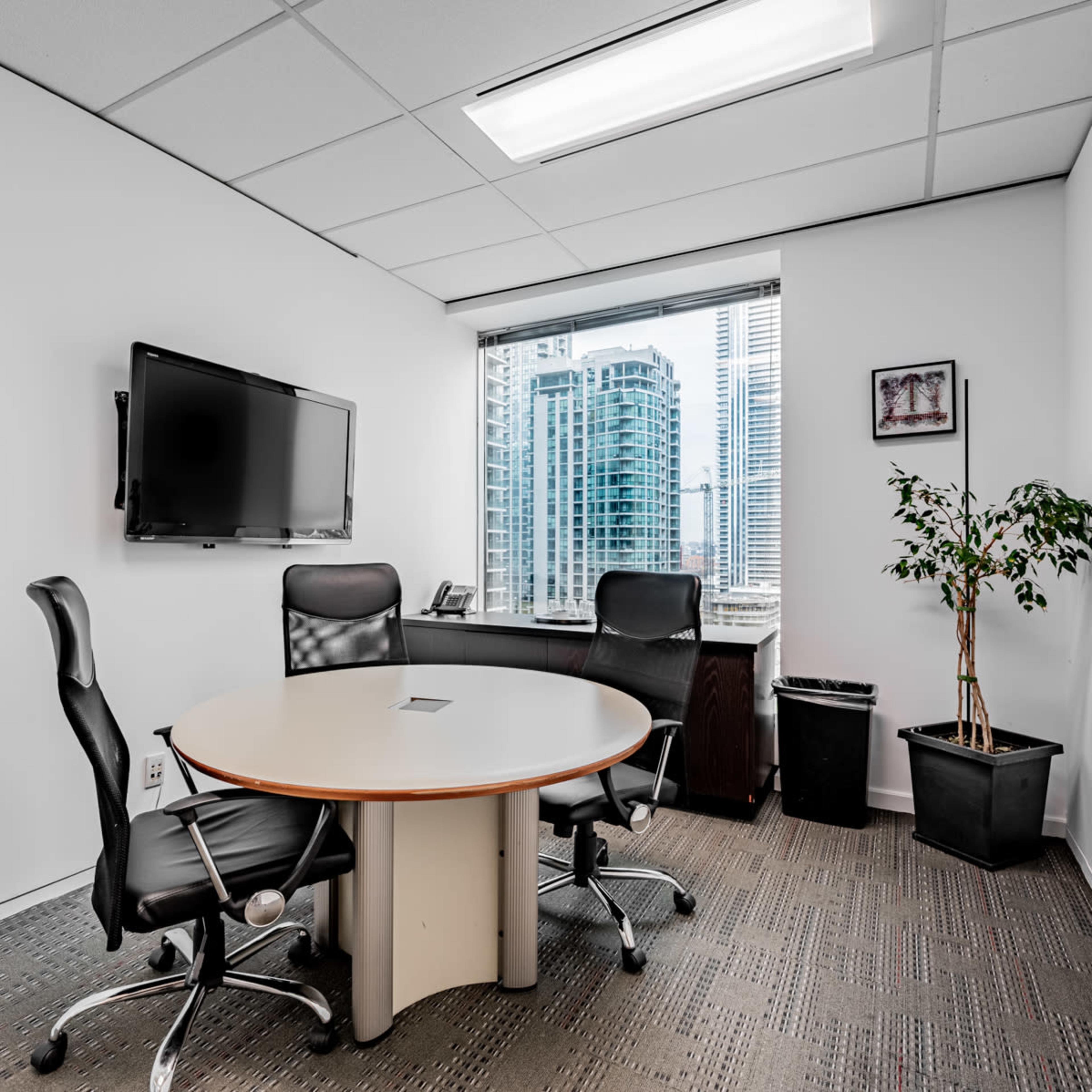 A modern meeting room with a round table, two black chairs, a wall-mounted TV, and a large window overlooking a cityscape.