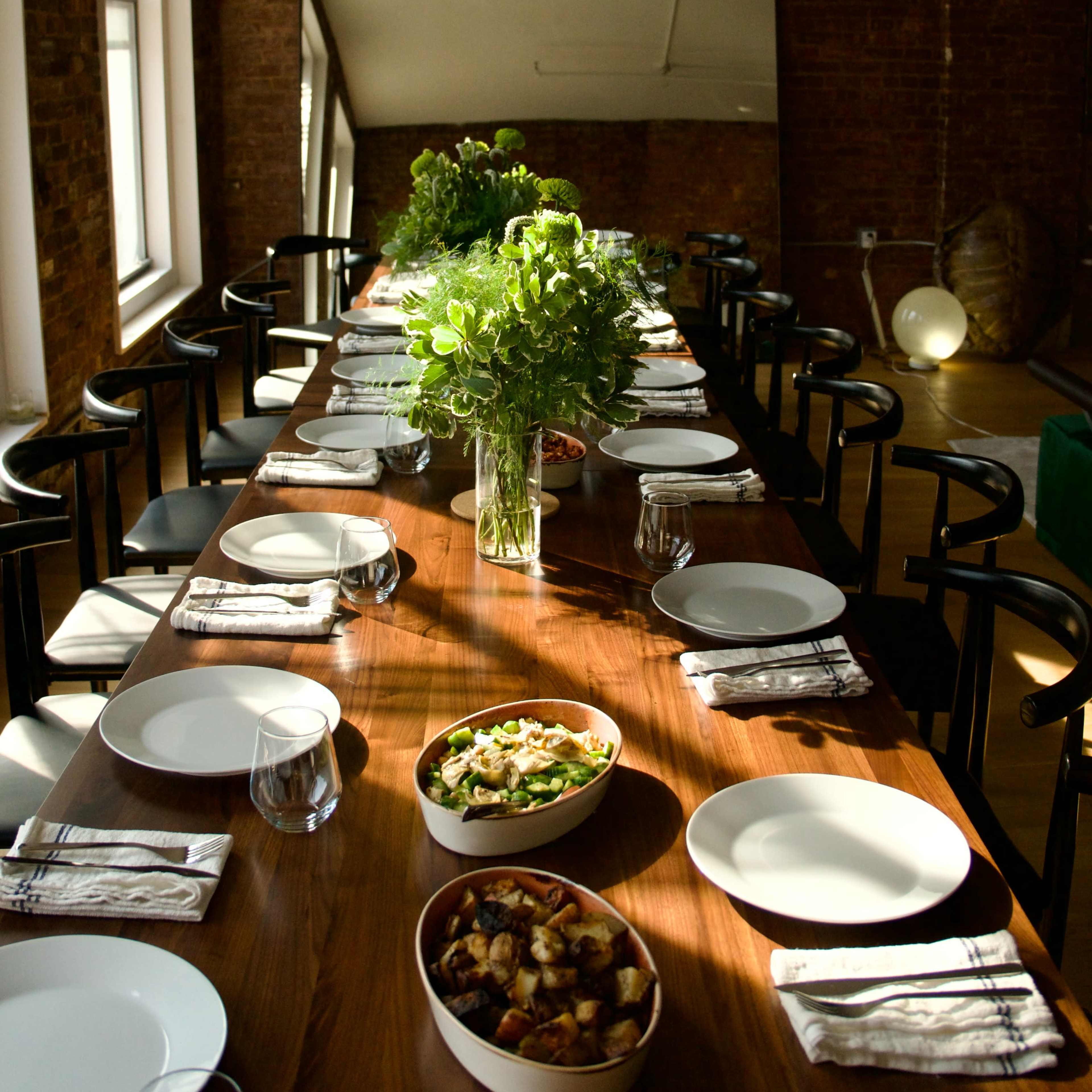 A long wooden dining table is set with white plates and silverware, featuring bowls of food in the center and surrounded by a simple floral arrangement.