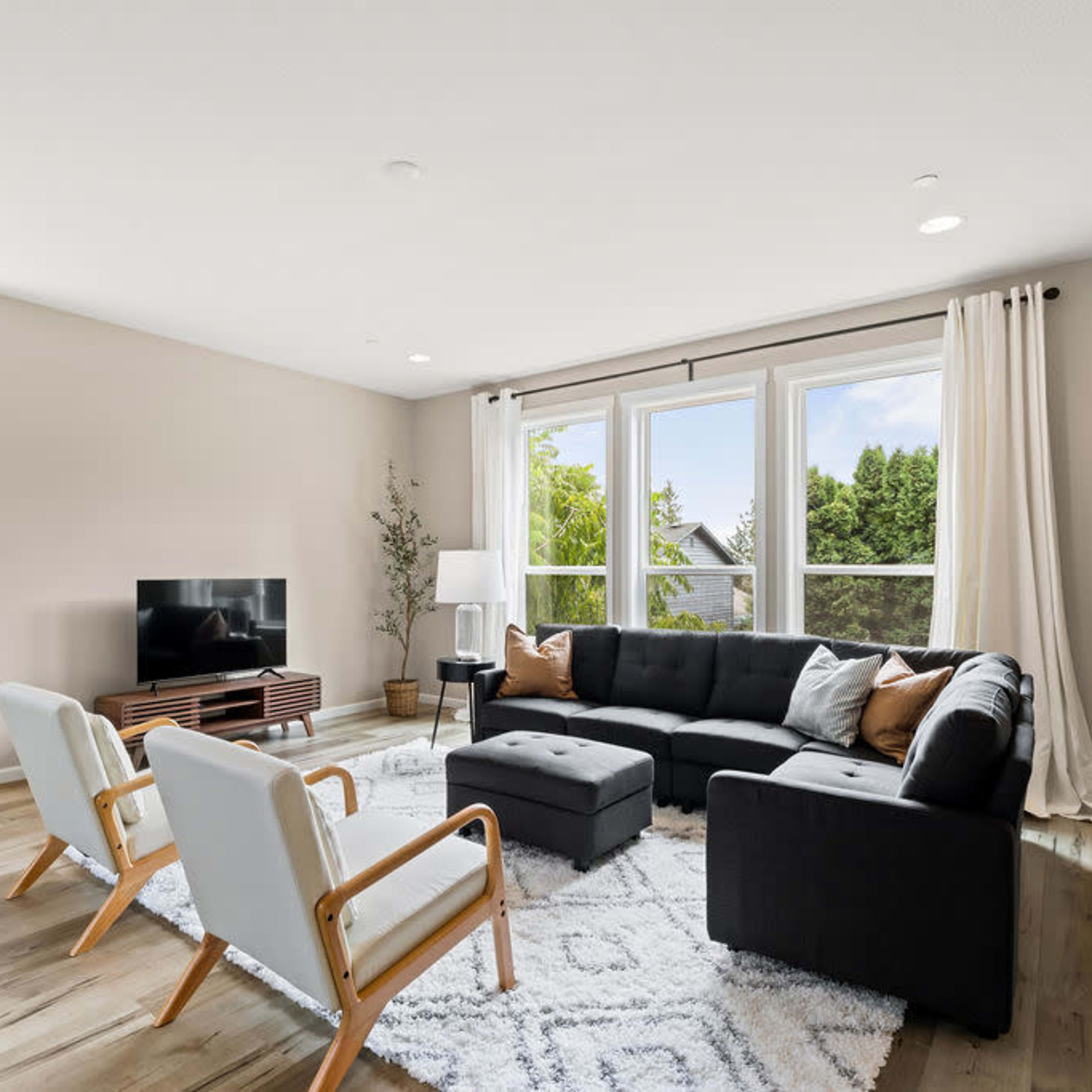 A modern living room features a black sectional sofa, two white chairs, a television on a wooden stand, and large windows overlooking greenery.