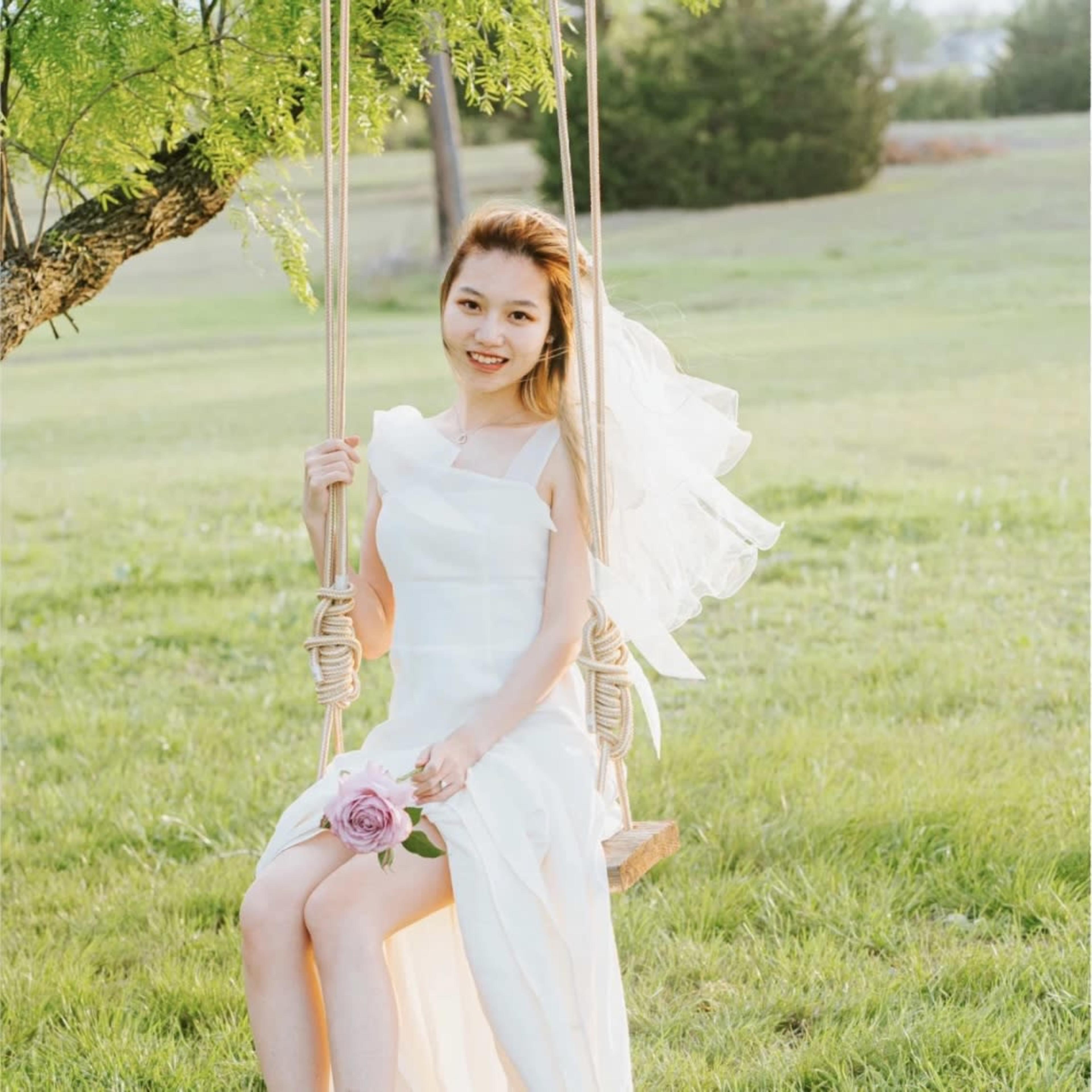 A woman in a white dress and veil sits on a swing under a tree, holding a rose, in an outdoor grassy setting.