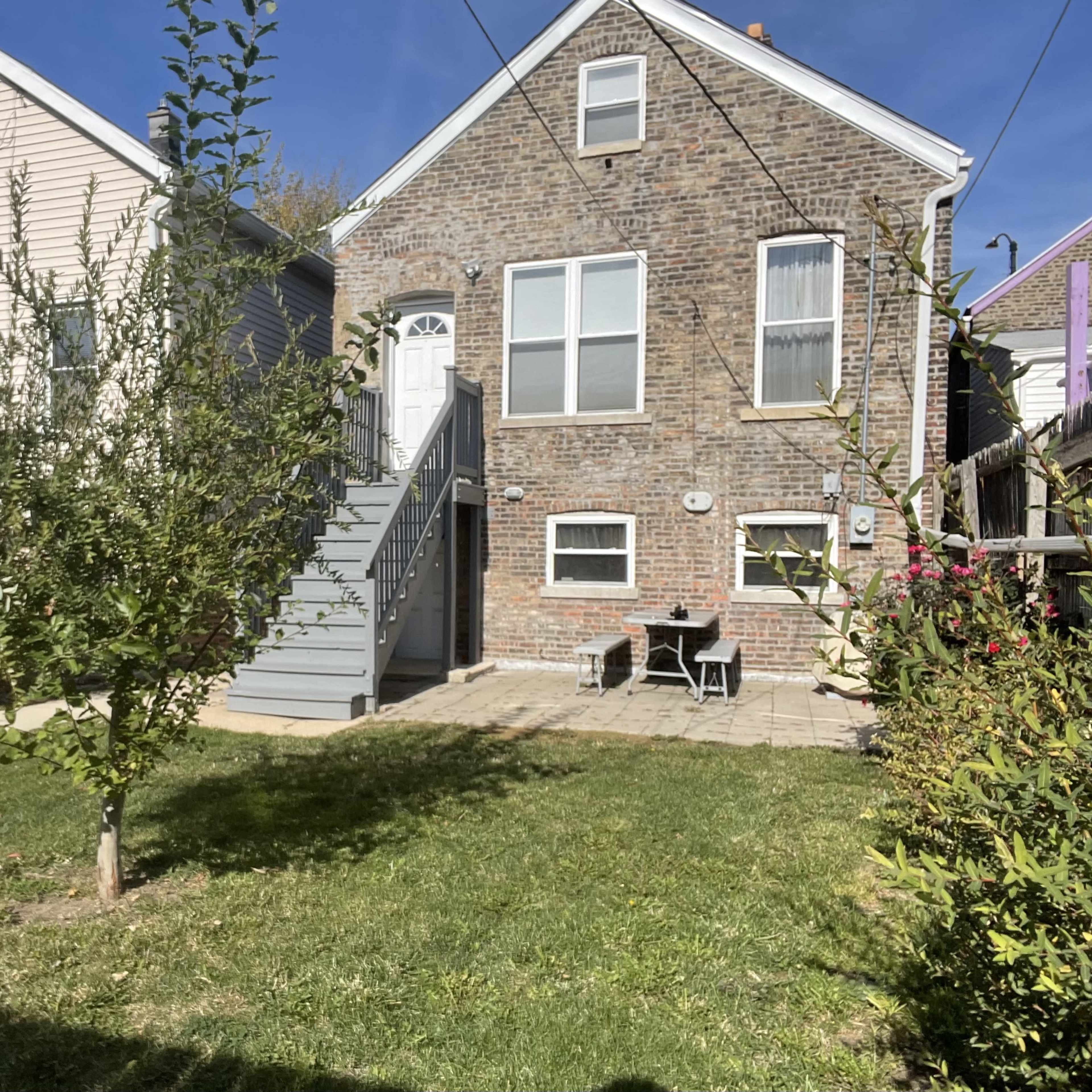 A residential yard with a brick house, a set of stairs leading to a front door, and patio furniture on a stone path surrounded by grass and a small tree.