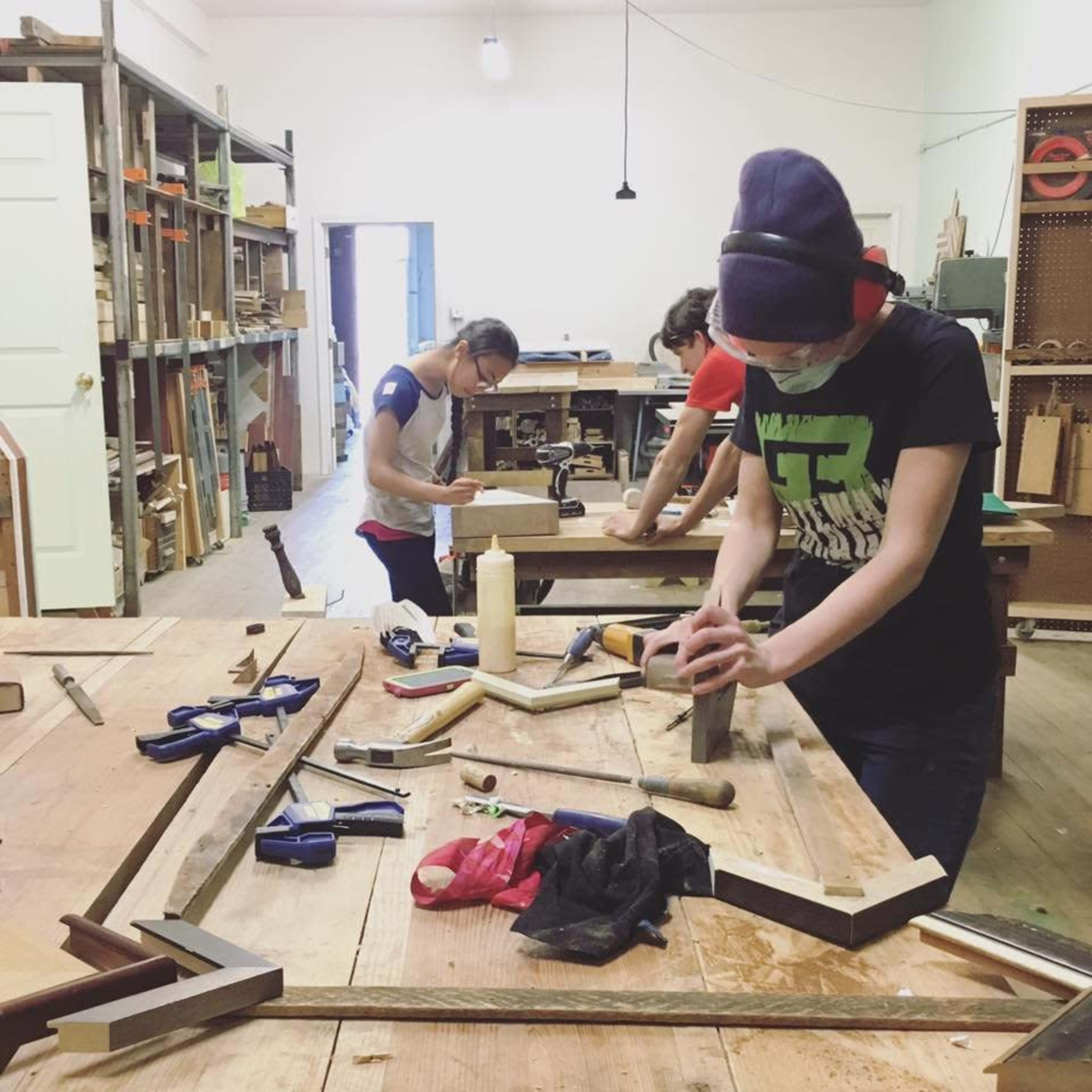 Three individuals work on woodworking projects in a workshop filled with tools and materials.