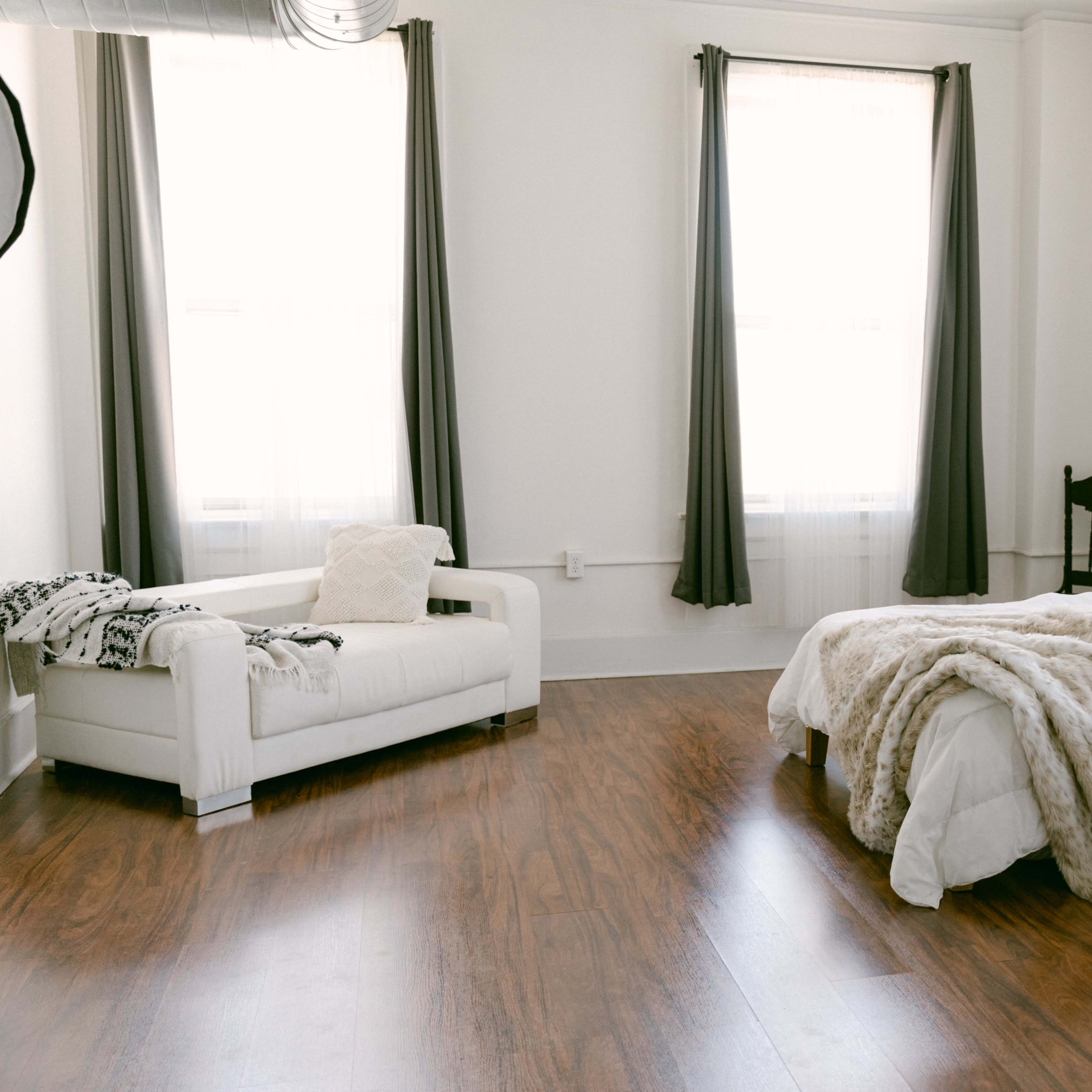 A bright and airy room with a bed dressed in white linens and a cozy throw, a white couch, and large windows dressed with grey curtains.