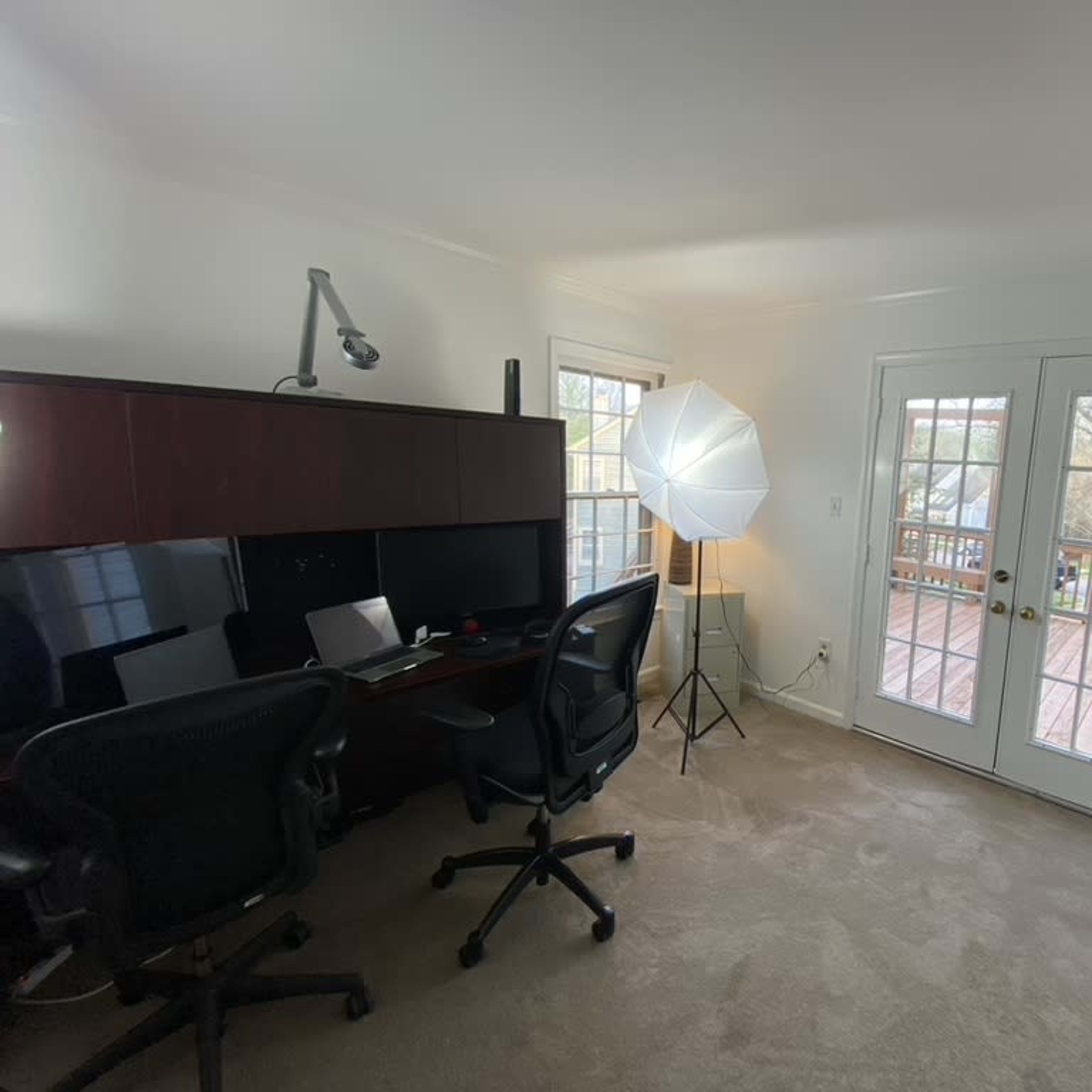 A well-lit office space with a wooden desk, two office chairs, and a softbox lighting setup next to a set of French doors leading to a deck.