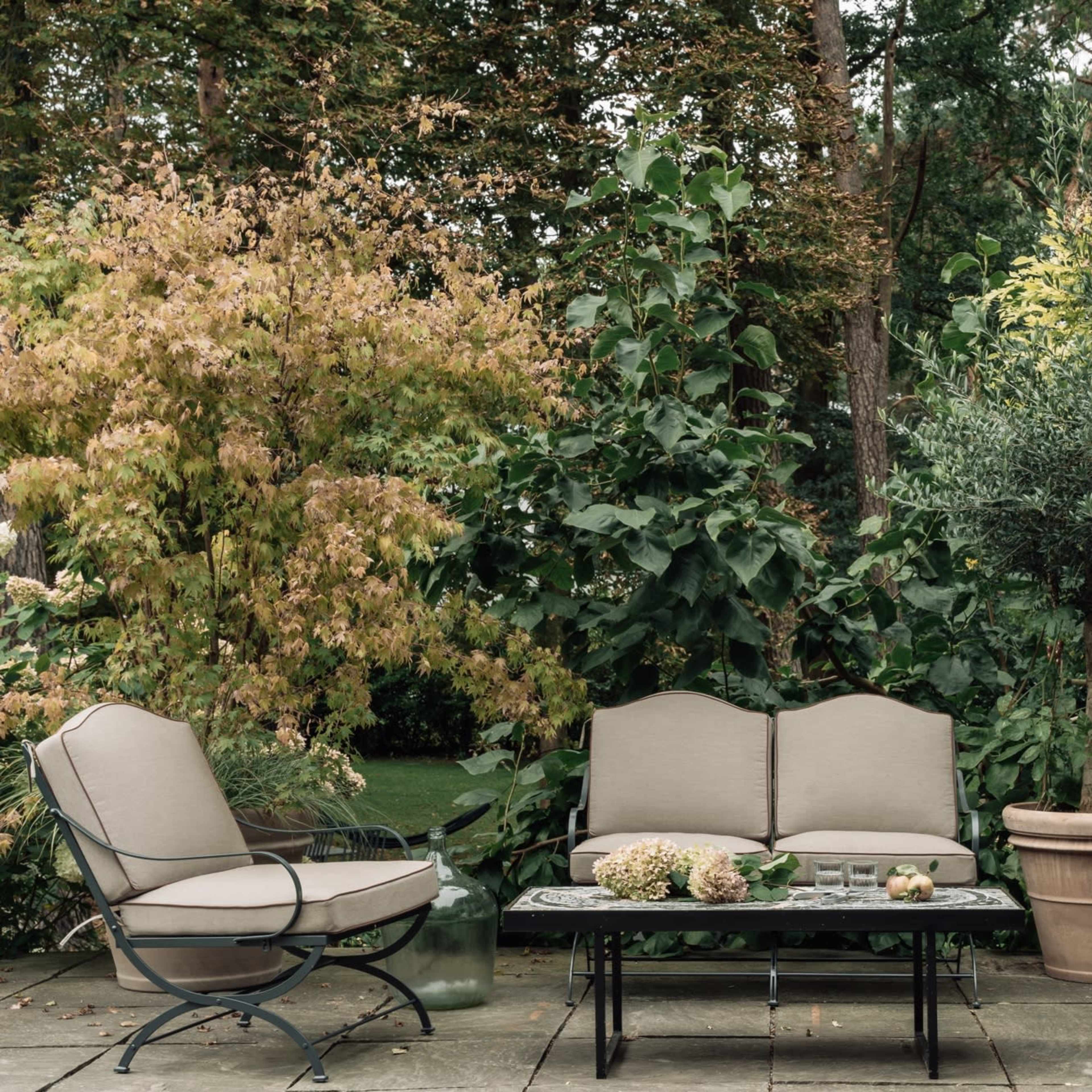 A seating area outdoors features three beige chairs and a coffee table surrounded by lush greenery and potted plants.