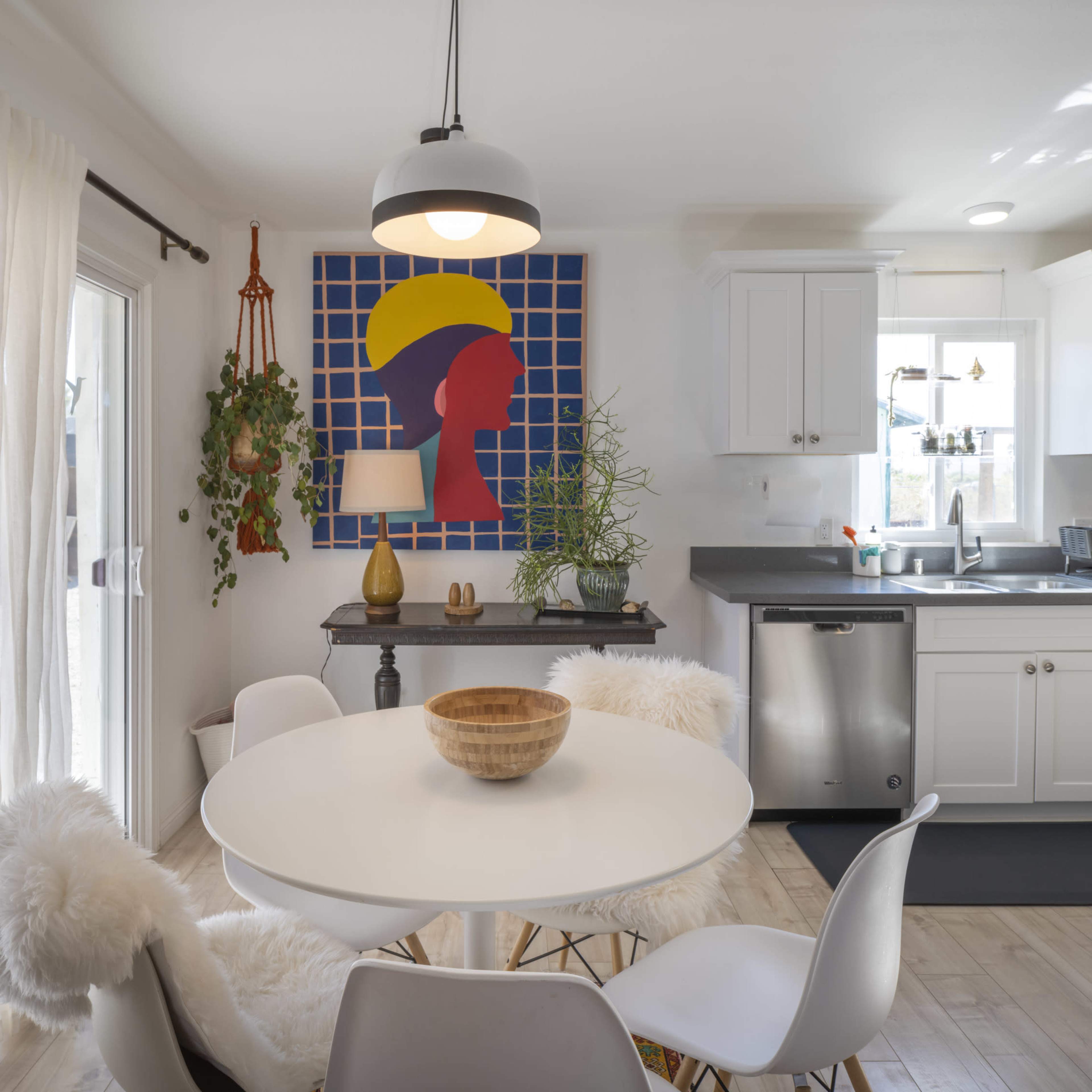 A bright kitchen features a round dining table with a wooden bowl, surrounded by white chairs, and is adorned with a colorful abstract wall art and plants.