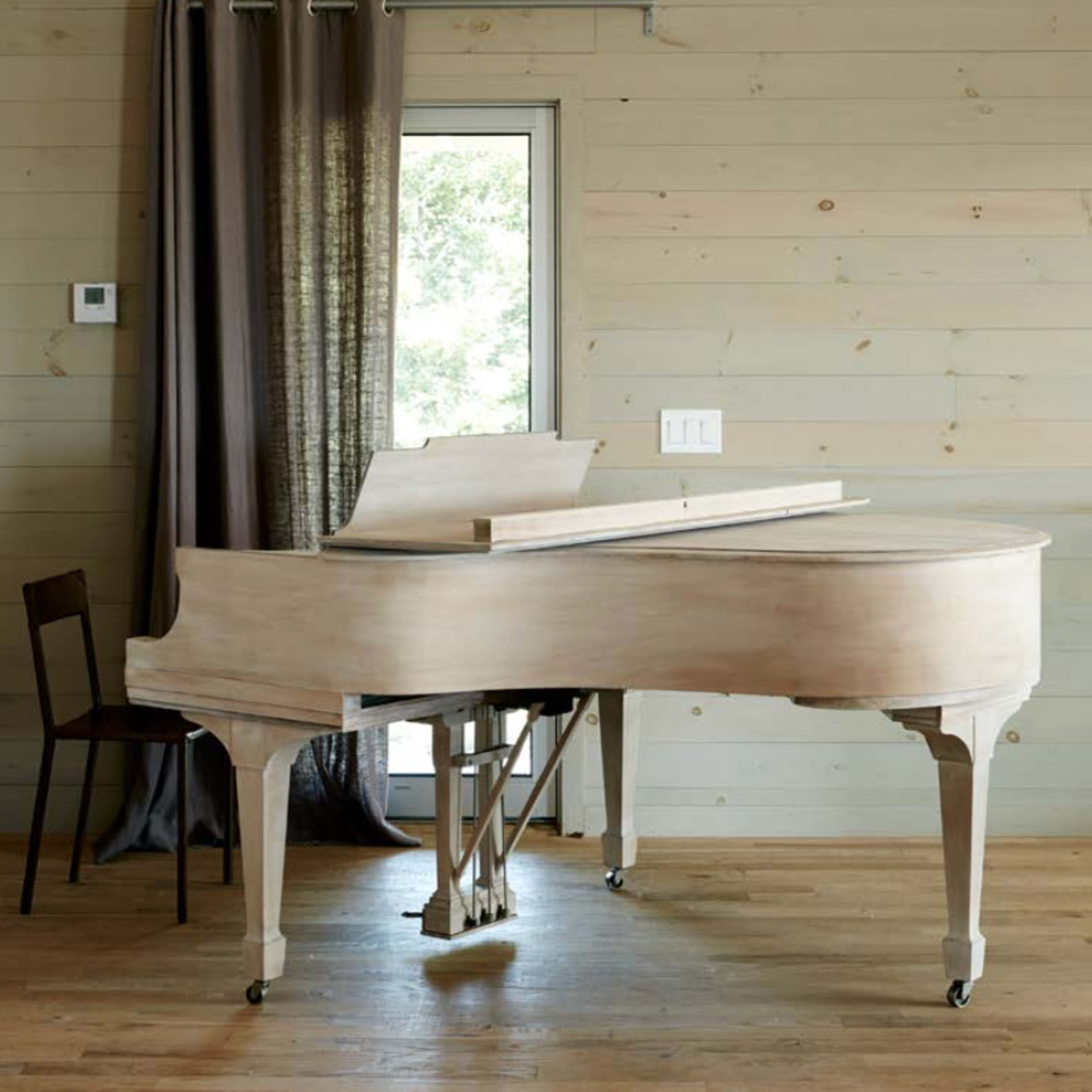 A wooden grand piano and a chair are positioned near a window in a room with wooden walls and floor.