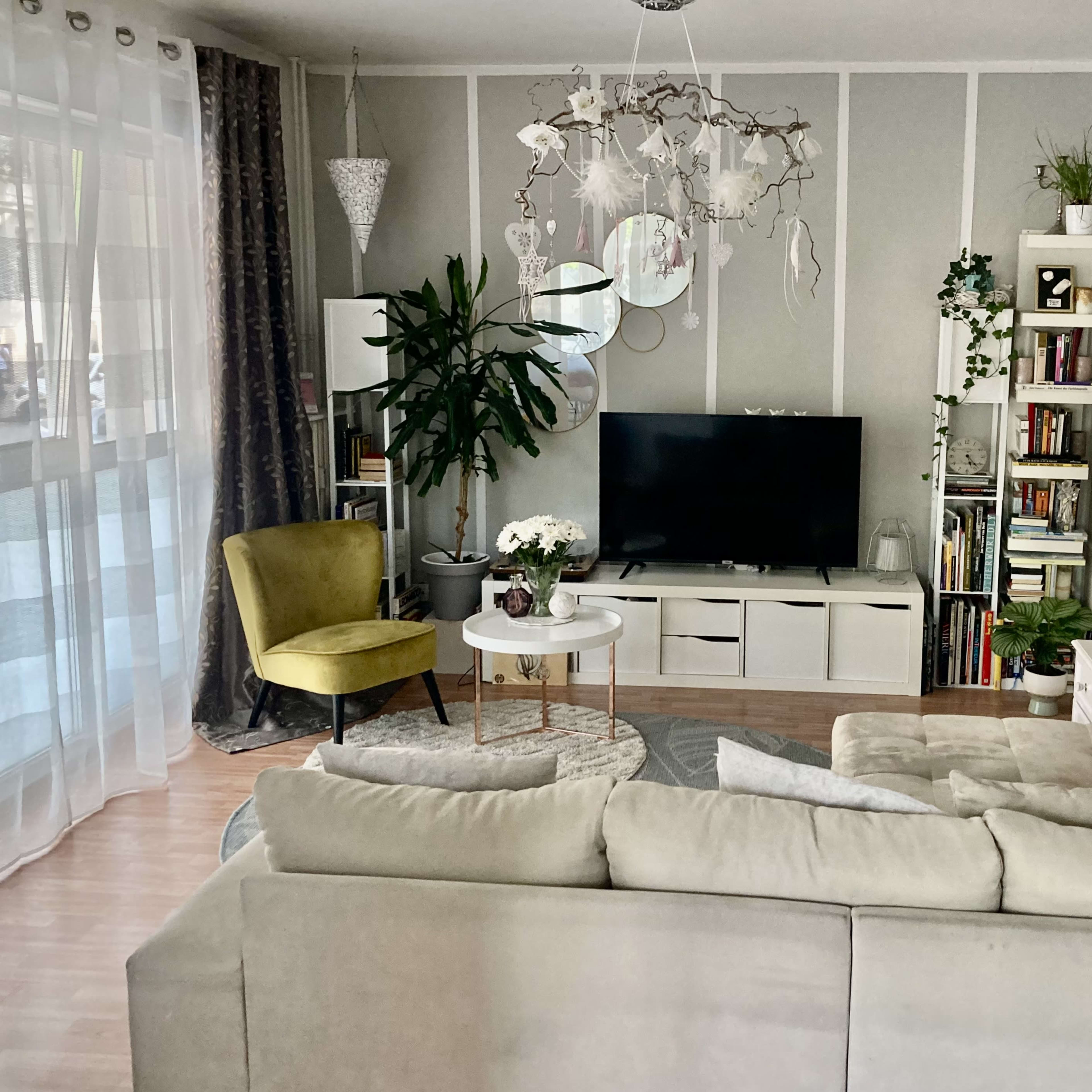 A living room with a beige sectional sofa, a green accent chair, a television on a white entertainment unit, and plants decorating the space.