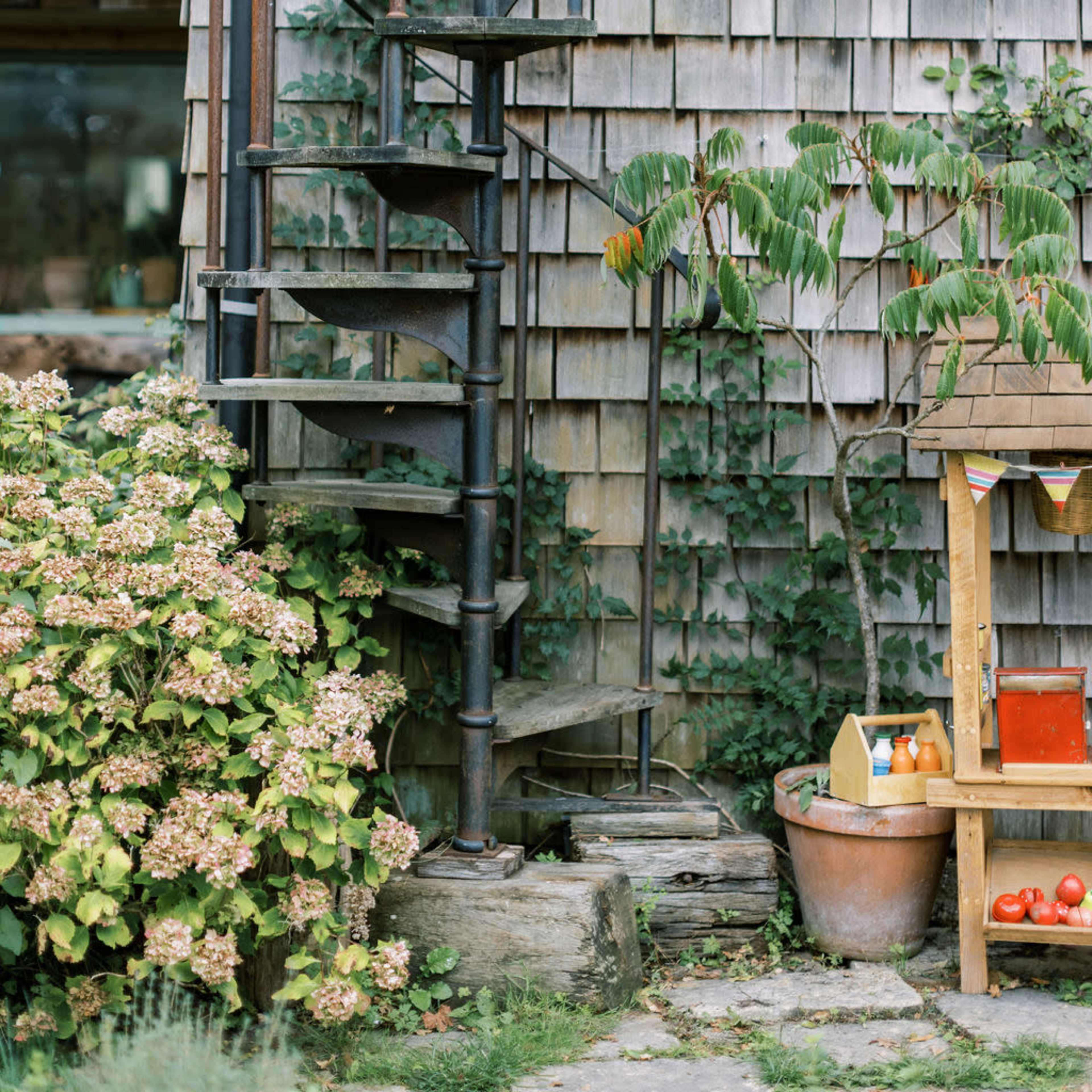 A metal spiral staircase rises alongside a wooden shingle exterior, surrounded by greenery and a wooden stand with various produce.