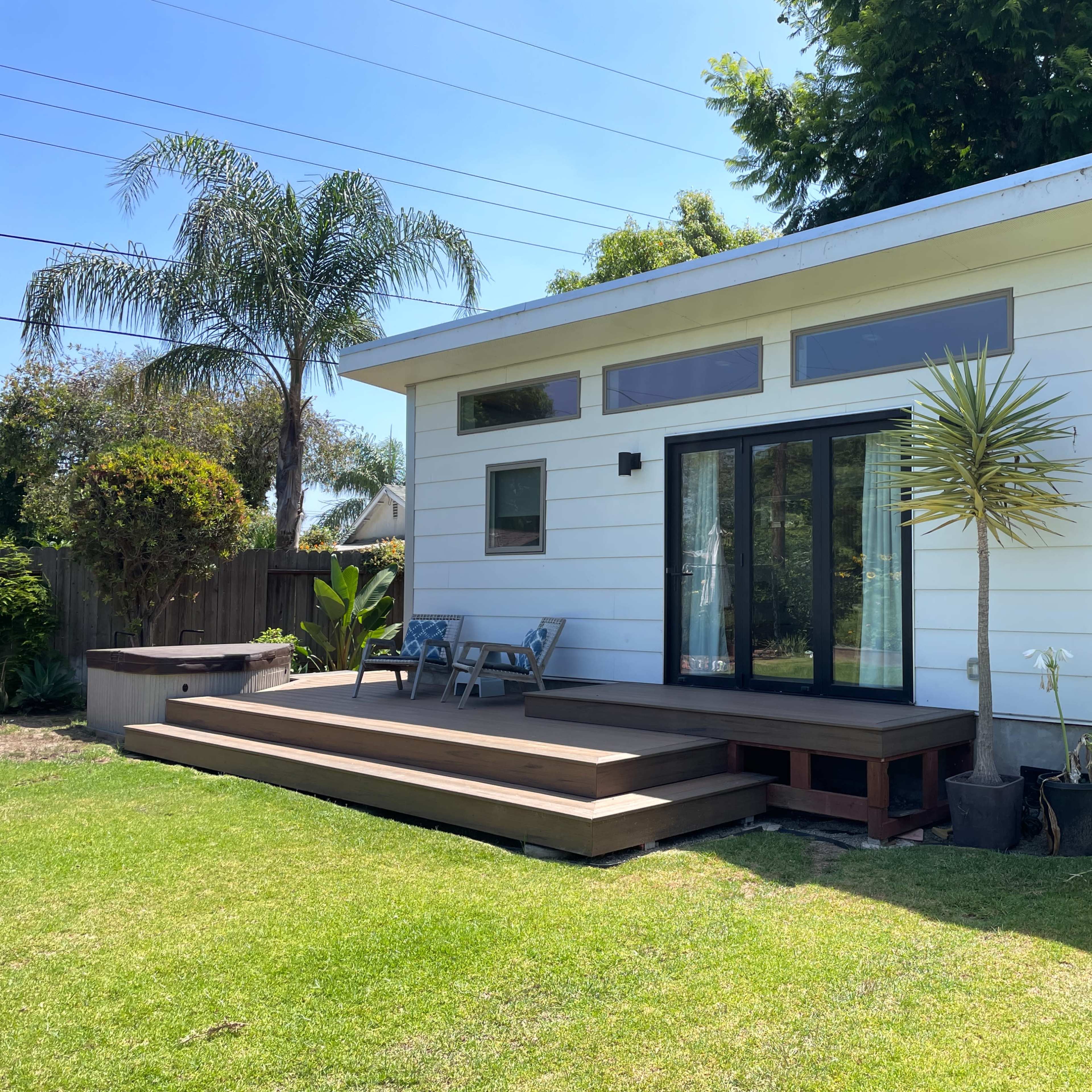 A small, modern white house features a wooden deck, large windows, and palm trees in the surrounding yard.