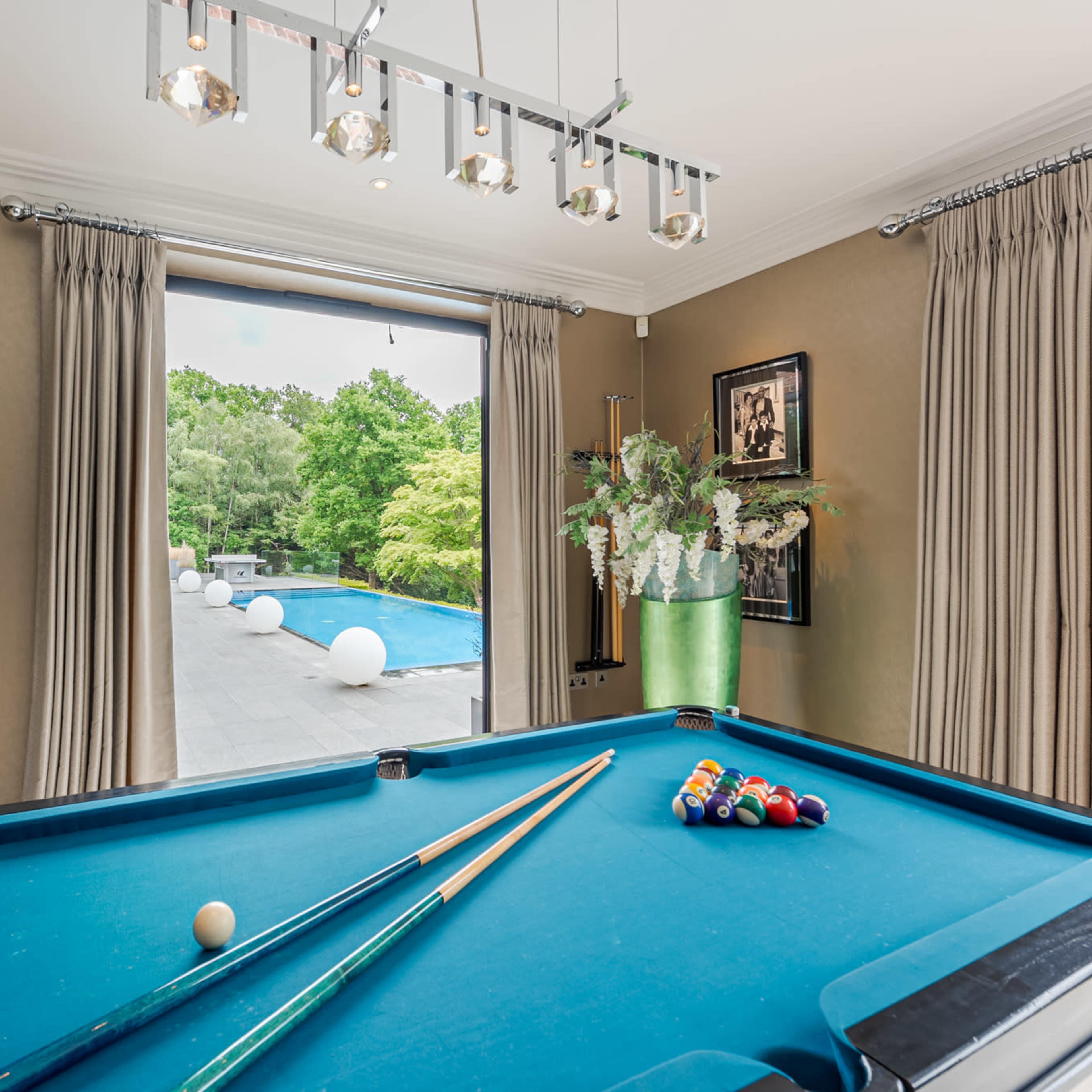 A pool table with cues and balls is positioned in a room with large windows overlooking a swimming pool and landscaped garden.