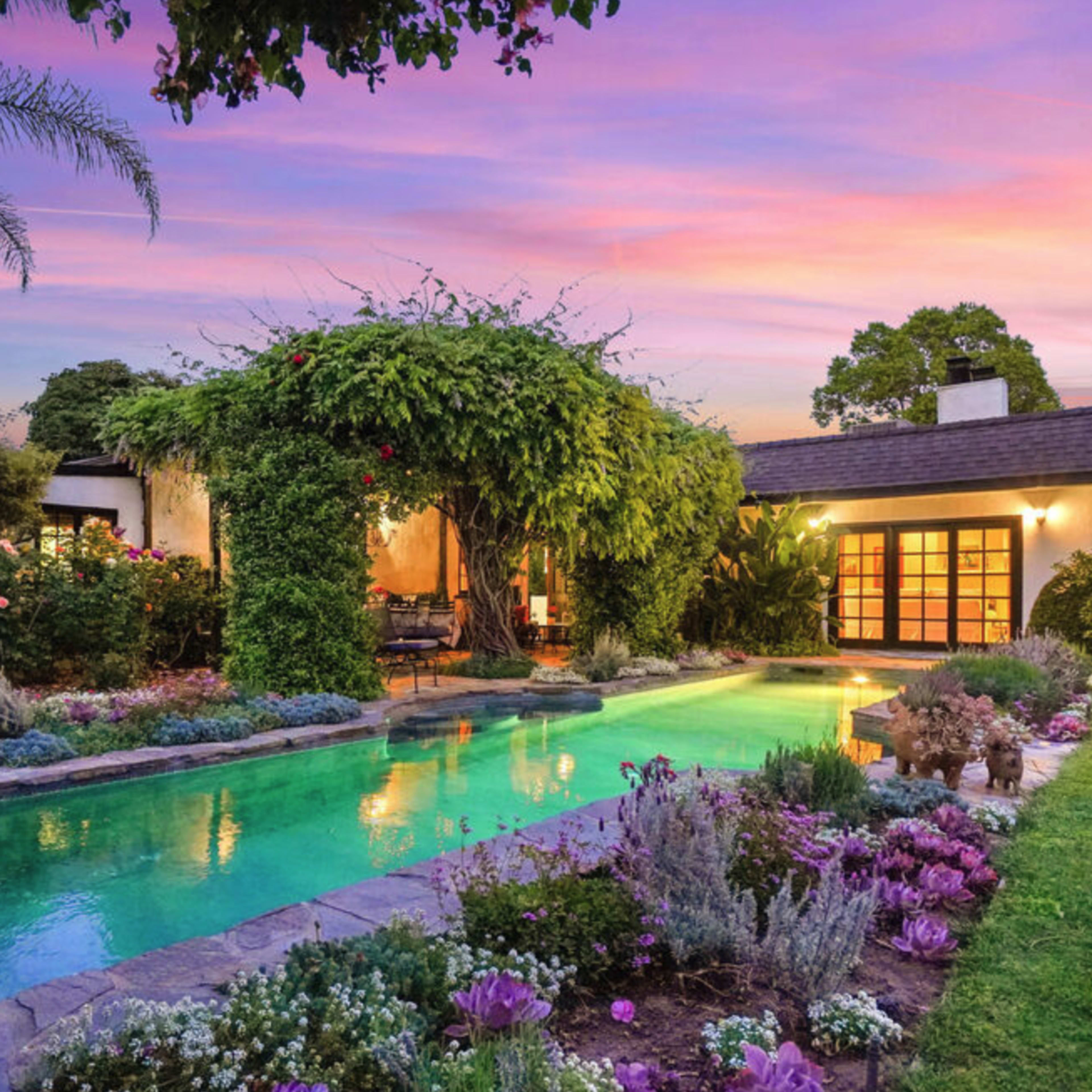 A landscaped backyard features a swimming pool surrounded by colorful flowers and greenery, with a house partially obscured by lush vines under a sunset sky.
