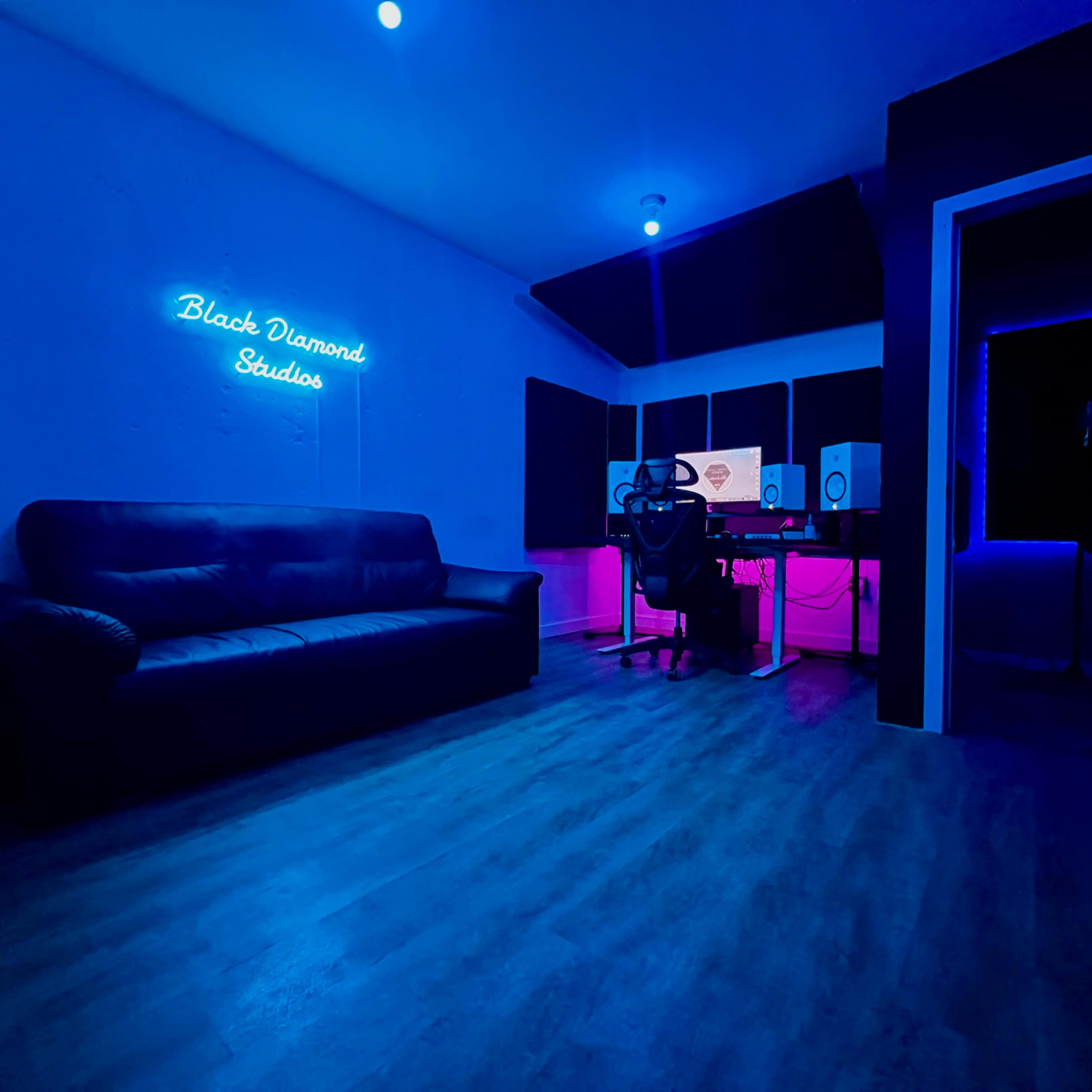 A dimly lit music studio with a black couch and a desk featuring audio equipment, illuminated by neon lighting.