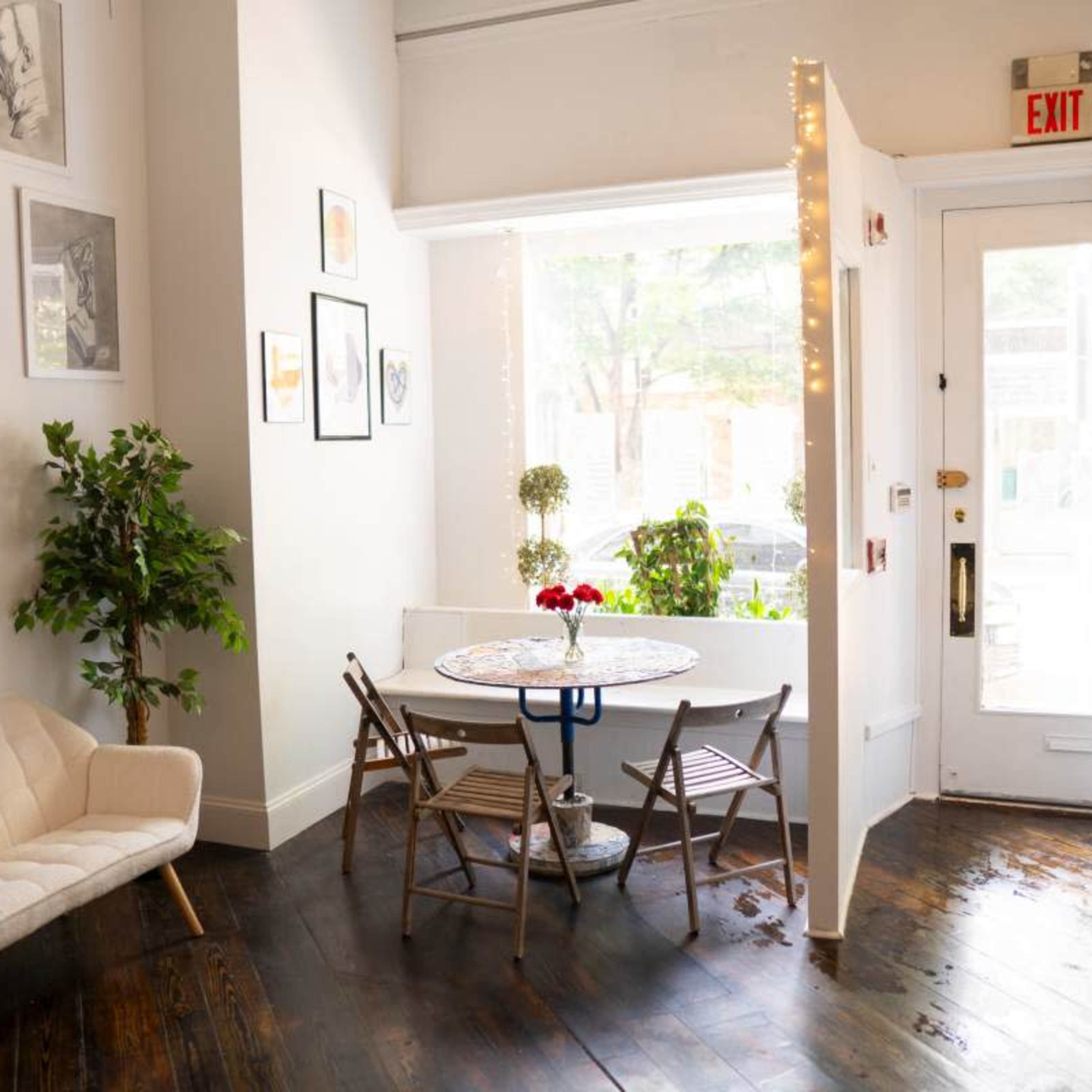A small café features a round table with chairs, a white couch, and large windows allowing natural light to fill the space.