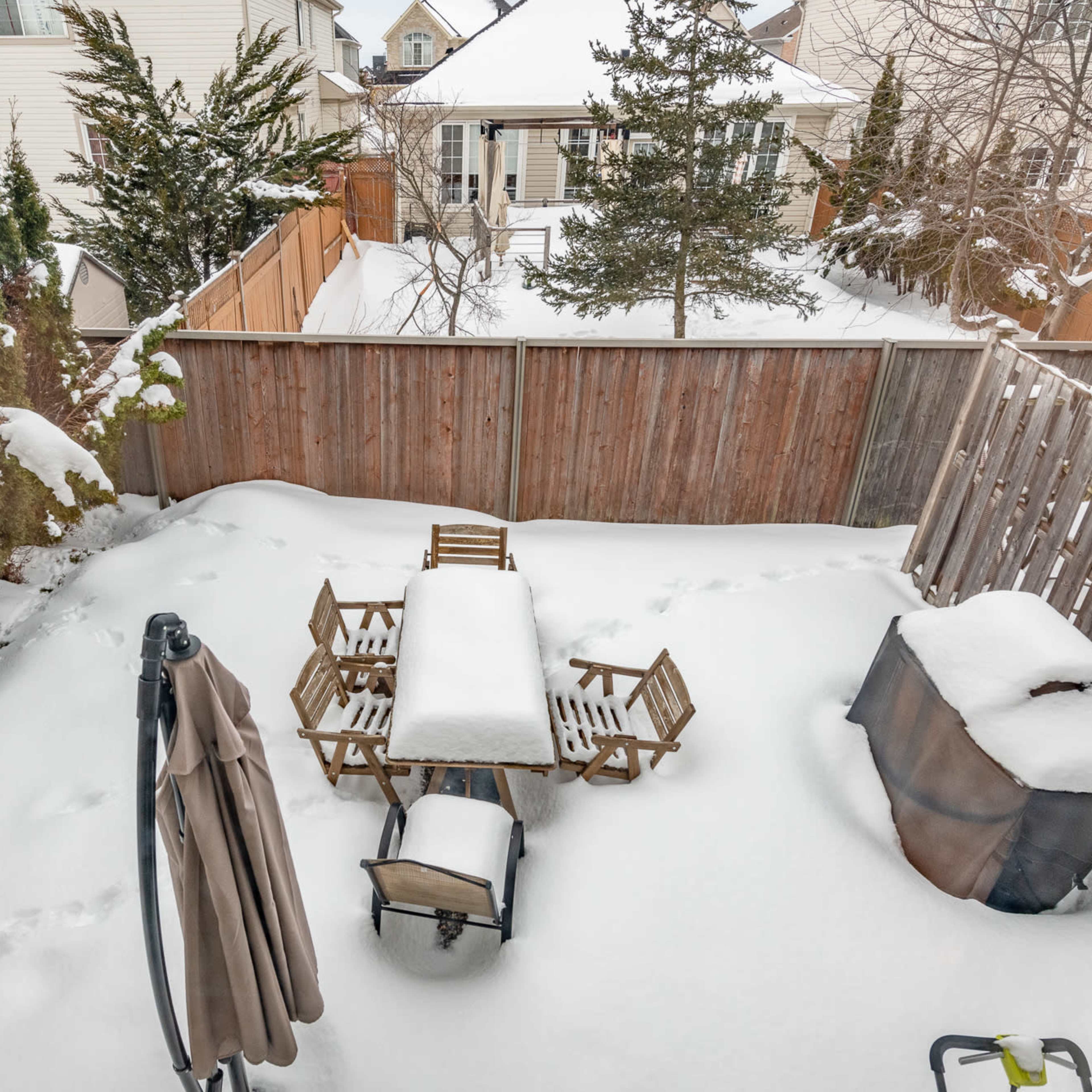 A backyard scene is covered in snow, featuring a table and chairs partially buried under a white blanket, with a grill and footprints visible in the snow.