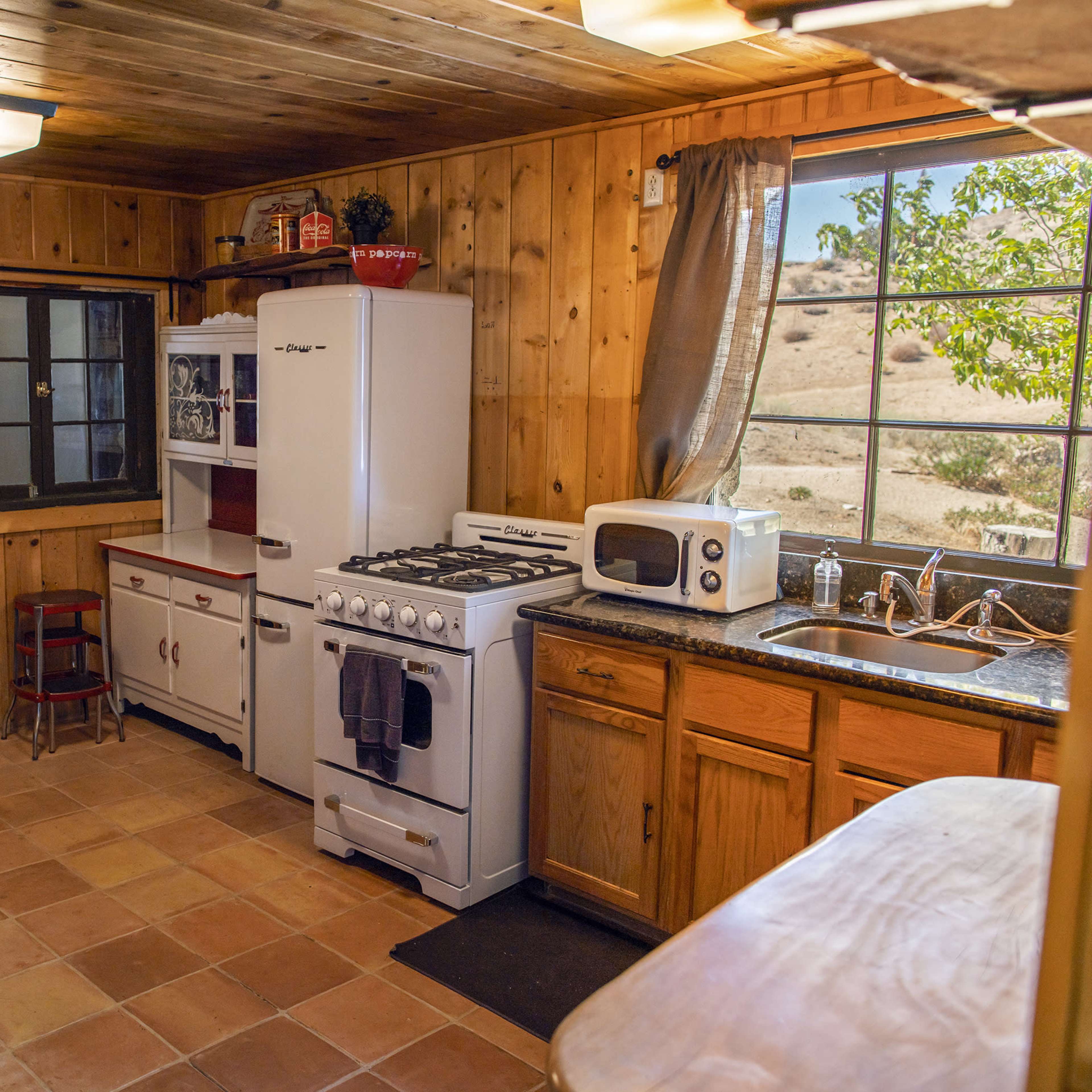 A rustic kitchen features wooden walls, a table, modern appliances, and a window overlooking a dry landscape.