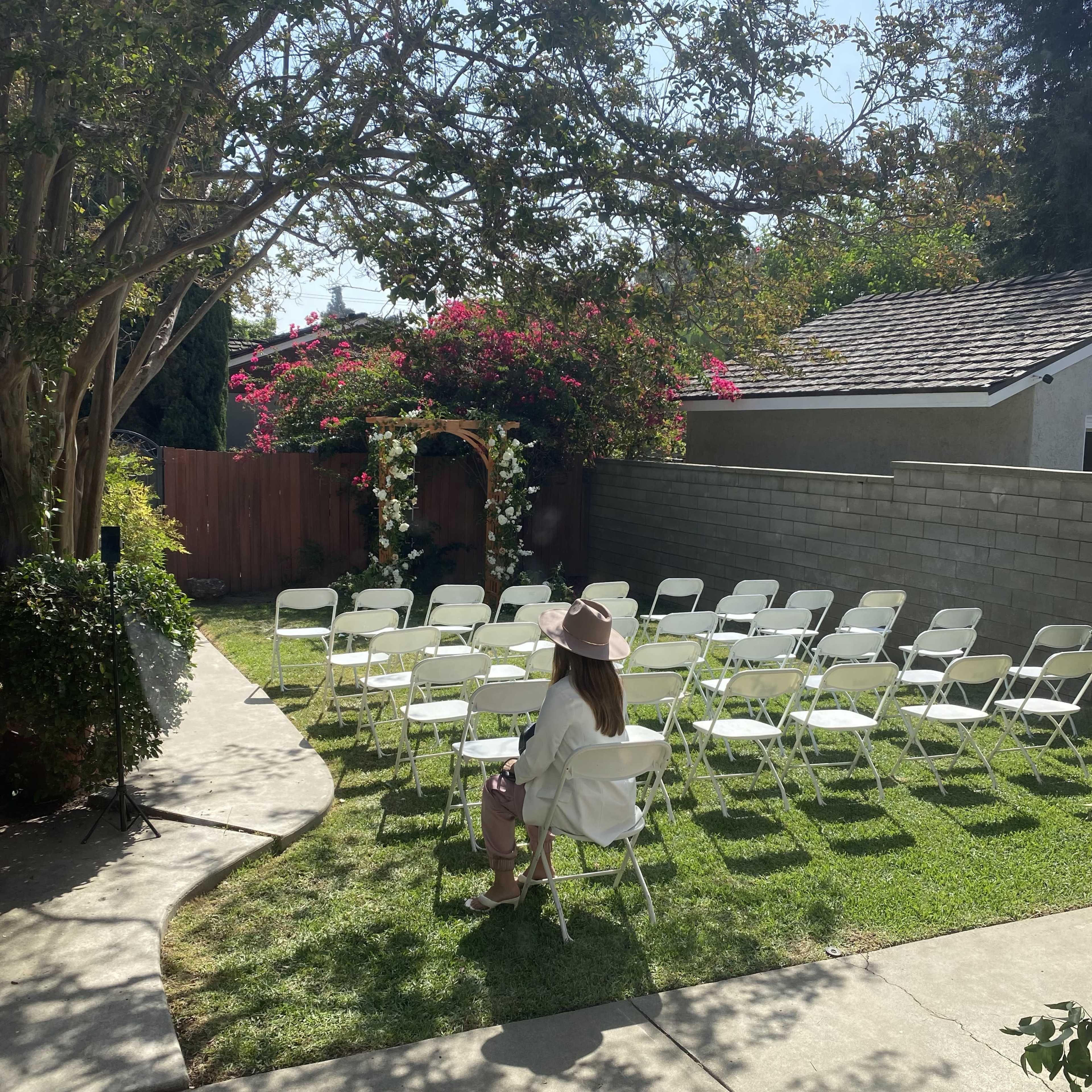 A woman sits alone in a white chair, facing a set of empty chairs arranged in a garden for an outdoor event.