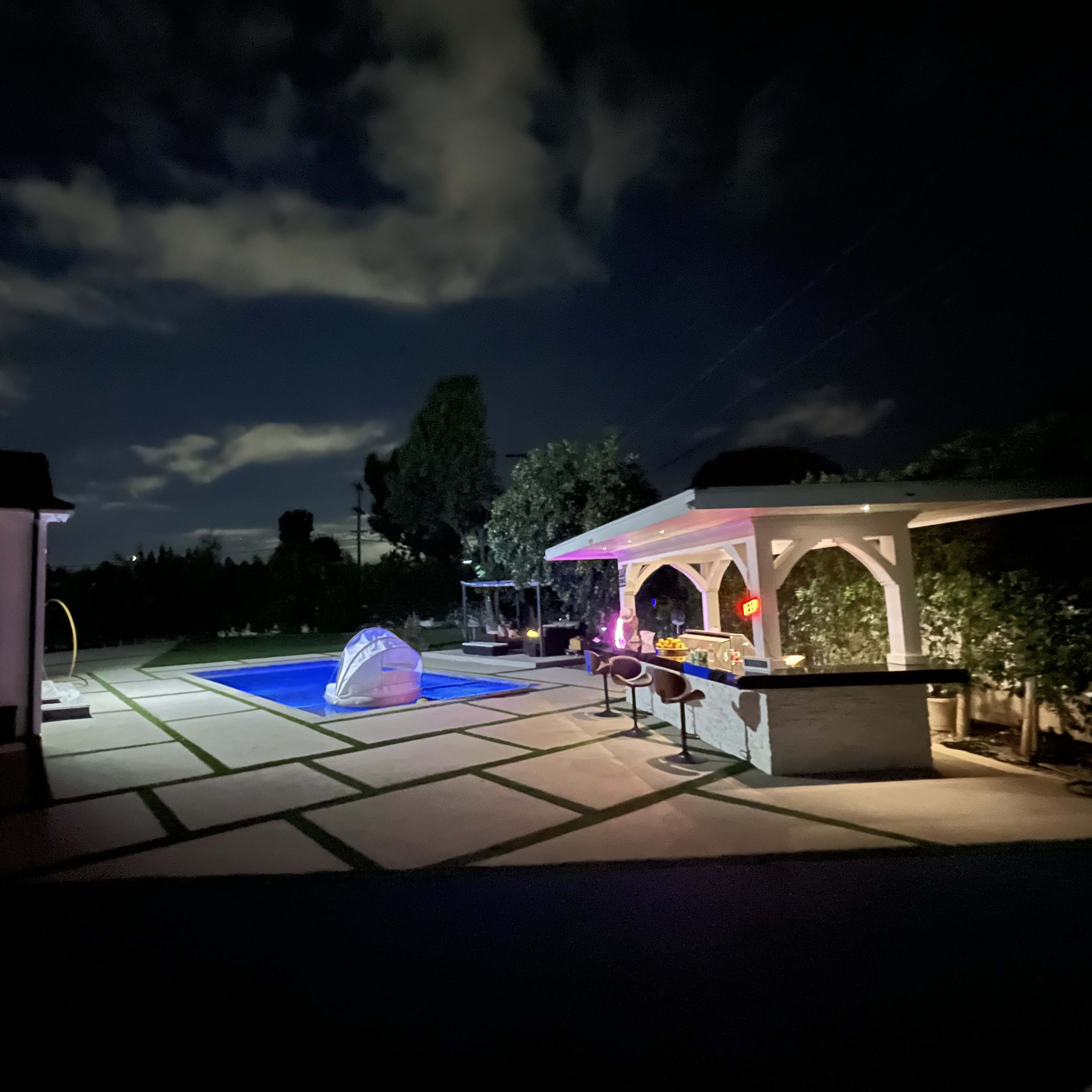 The scene features a well-lit backyard at night with a swimming pool, a bar area, and cloudy skies.