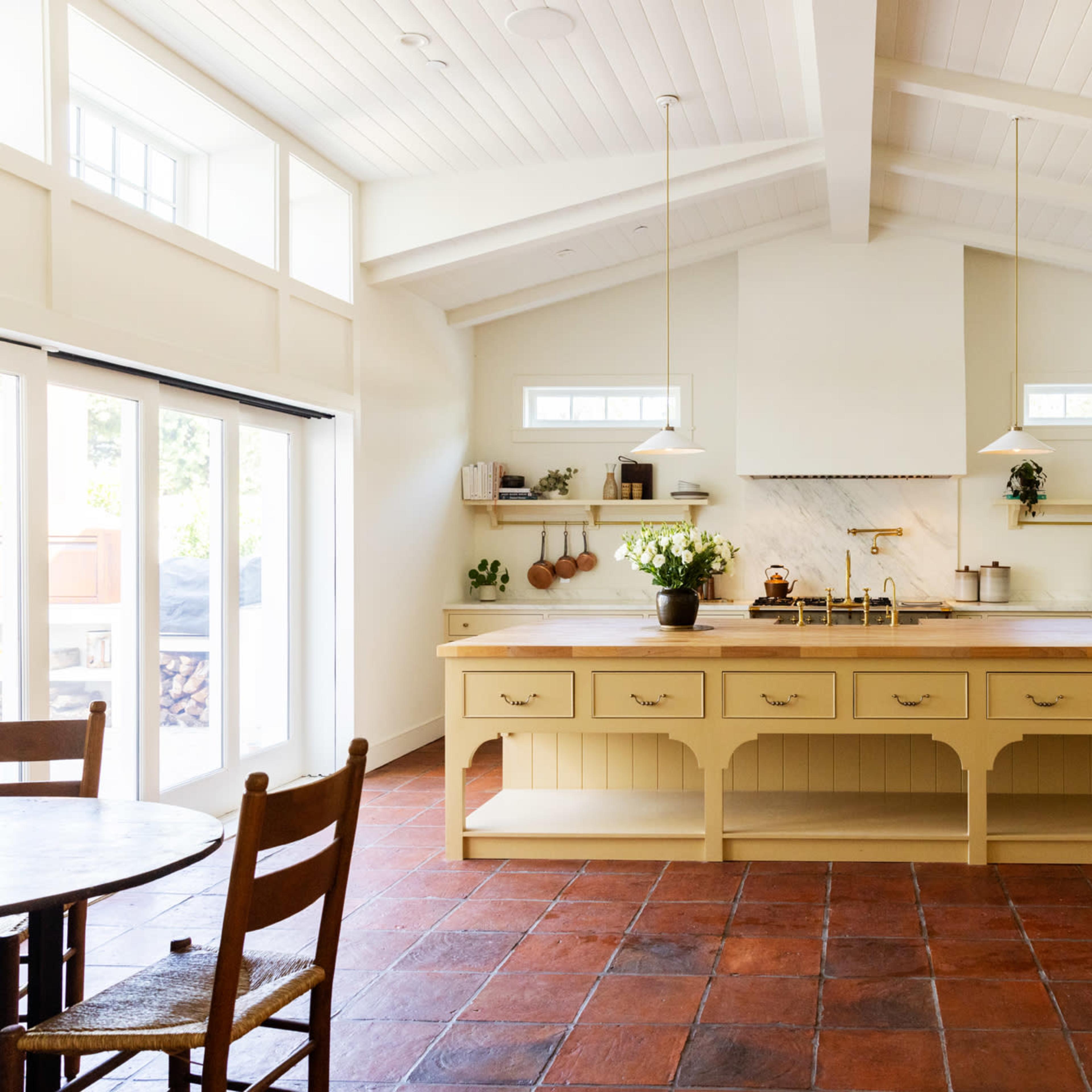 A spacious kitchen features a large central island with bar stools, white cabinetry, and terracotta tiled flooring.