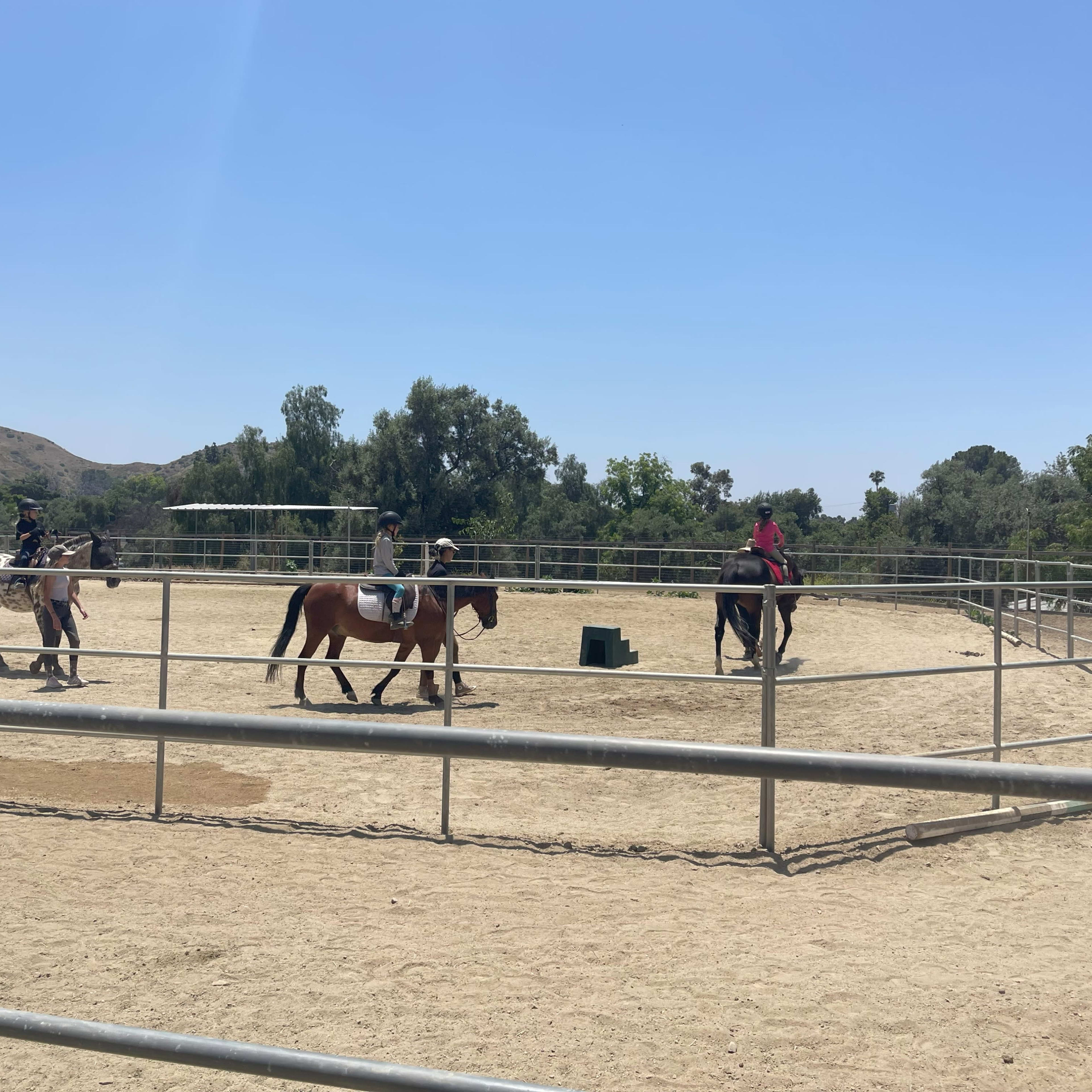Two riders are on horseback in a sandy arena surrounded by trees and hills under a clear sky.