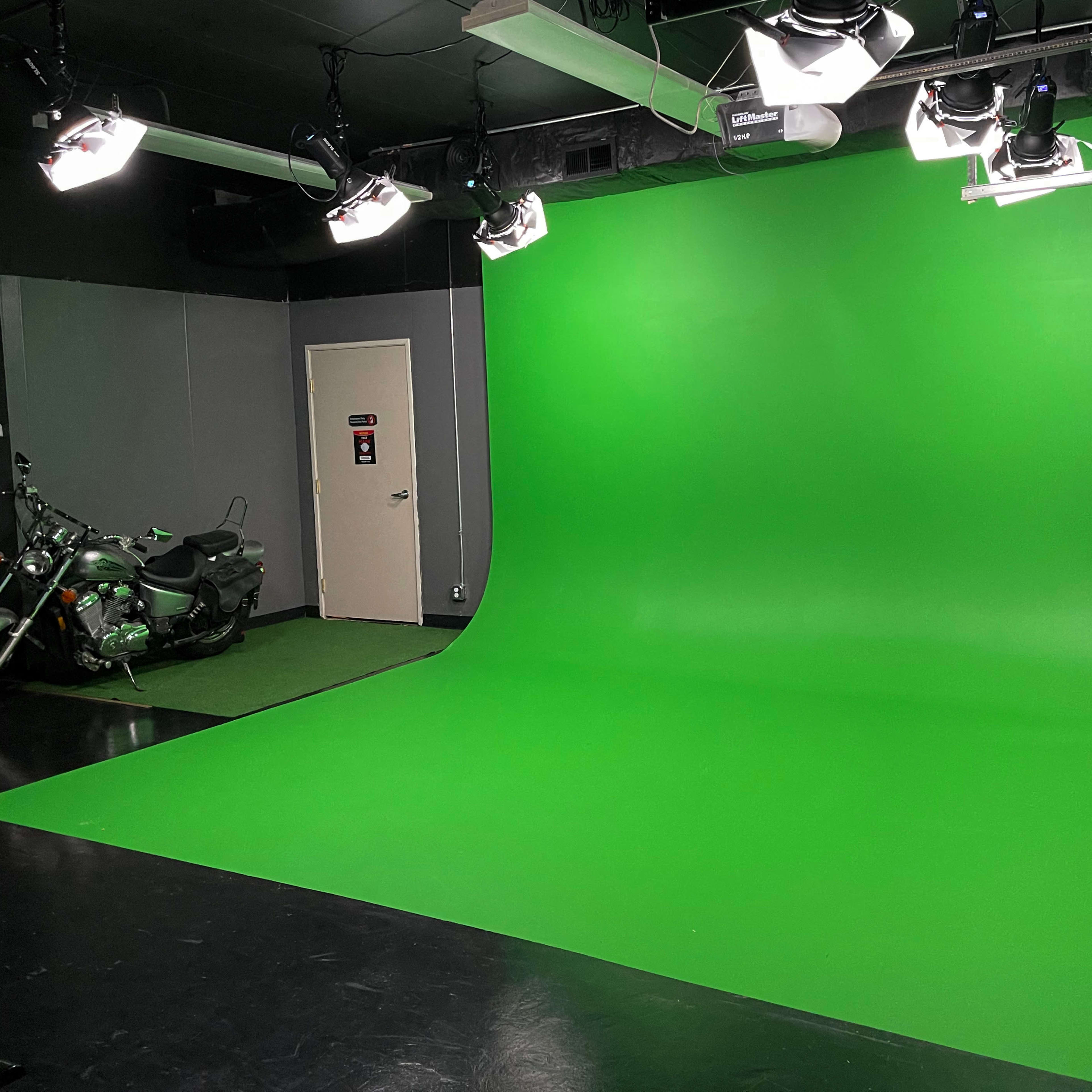 A studio space featuring a large green screen background, equipped with lighting fixtures and a motorcycle positioned on the floor.