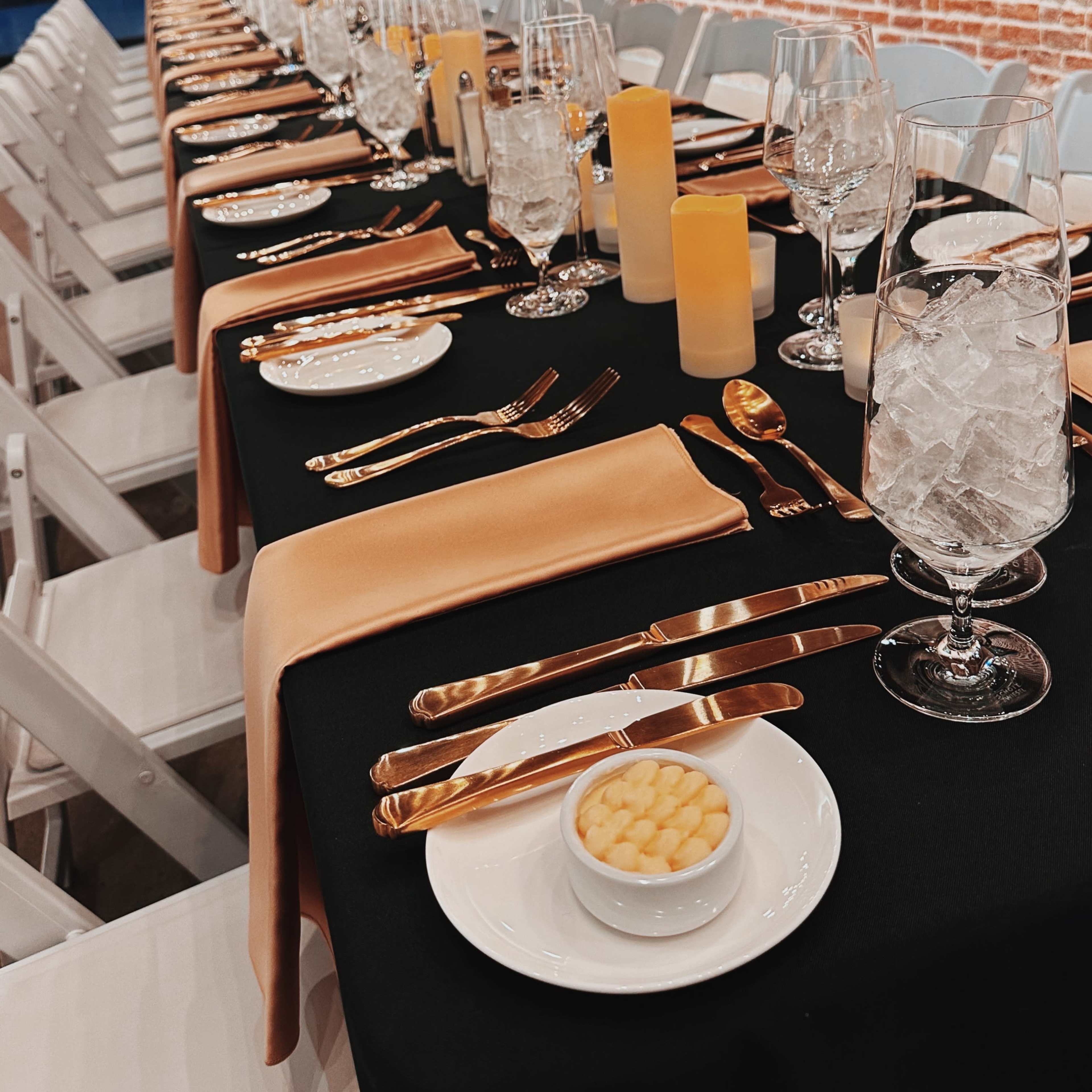 A long dining table is set with elegant dishes, glassware, and golden utensils, adorned with candles and decorative artwork in a rustic space.