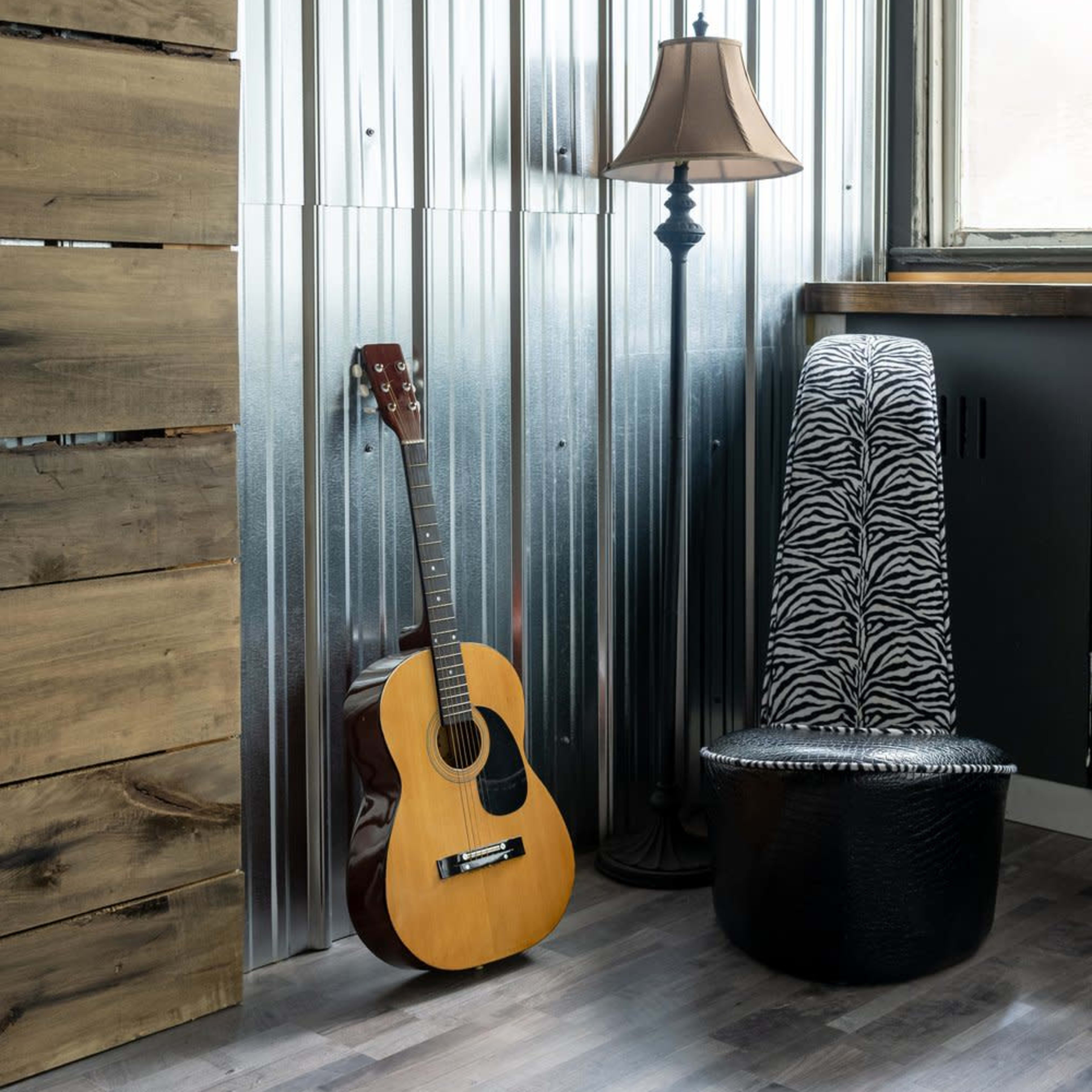 A wooden acoustic guitar leans against a metallic wall near a lamp and a zebra-patterned chair in a room.