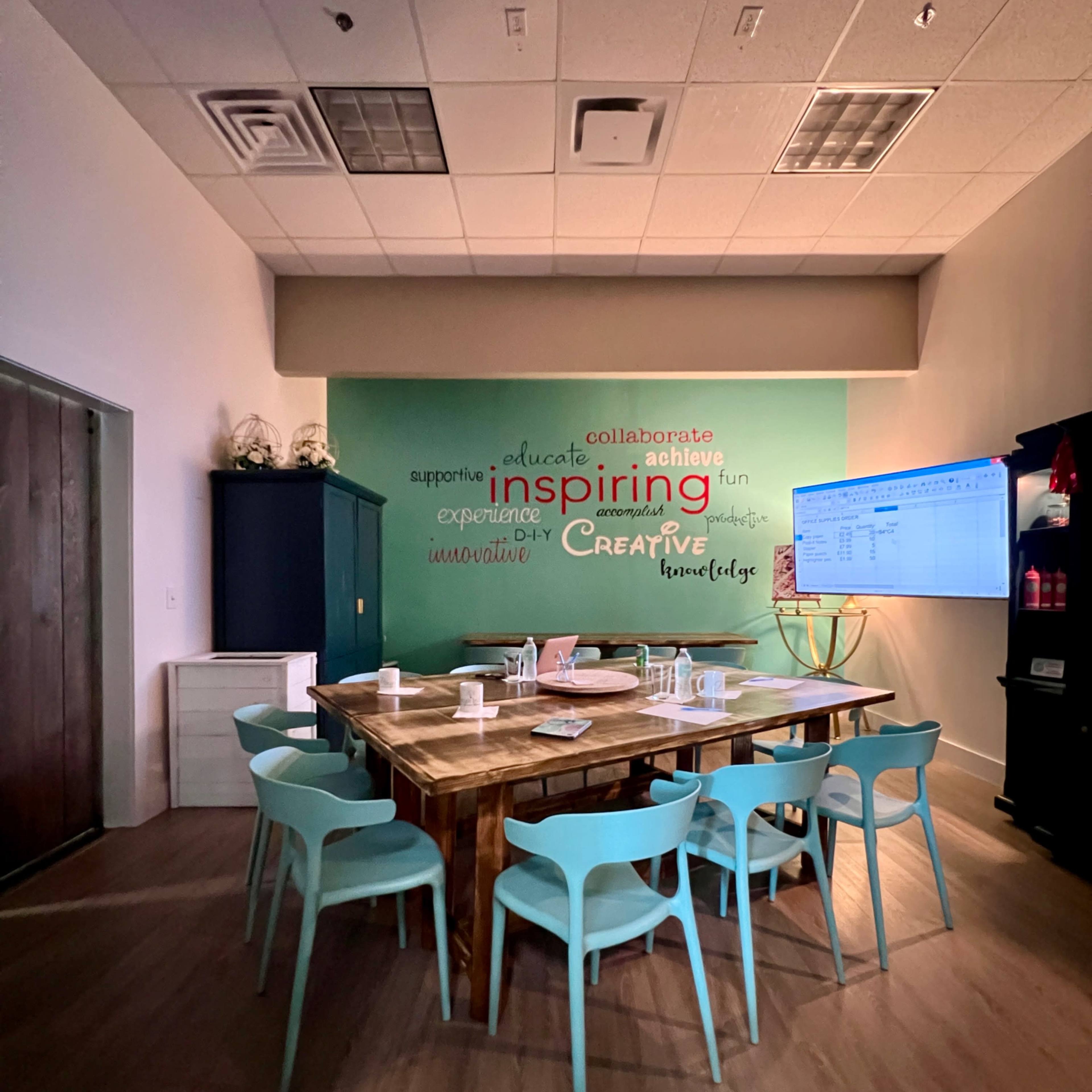 A meeting room features a large wooden table surrounded by light blue chairs, with a green wall displaying motivational words and a screen on one side.