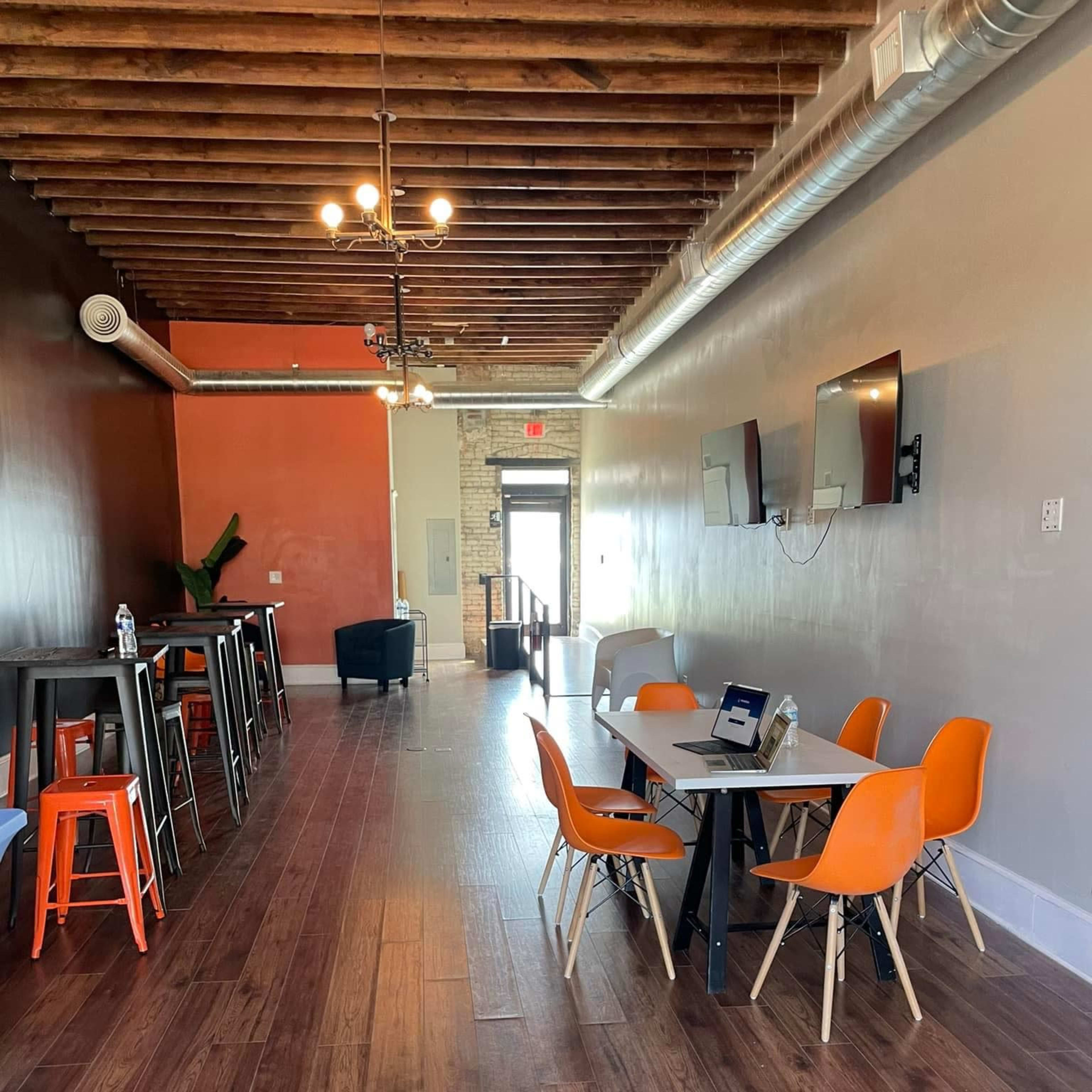 A modern, open interior space featuring wooden floors, a long table with orange and white chairs, a bar area with high stools, and two wall-mounted televisions.