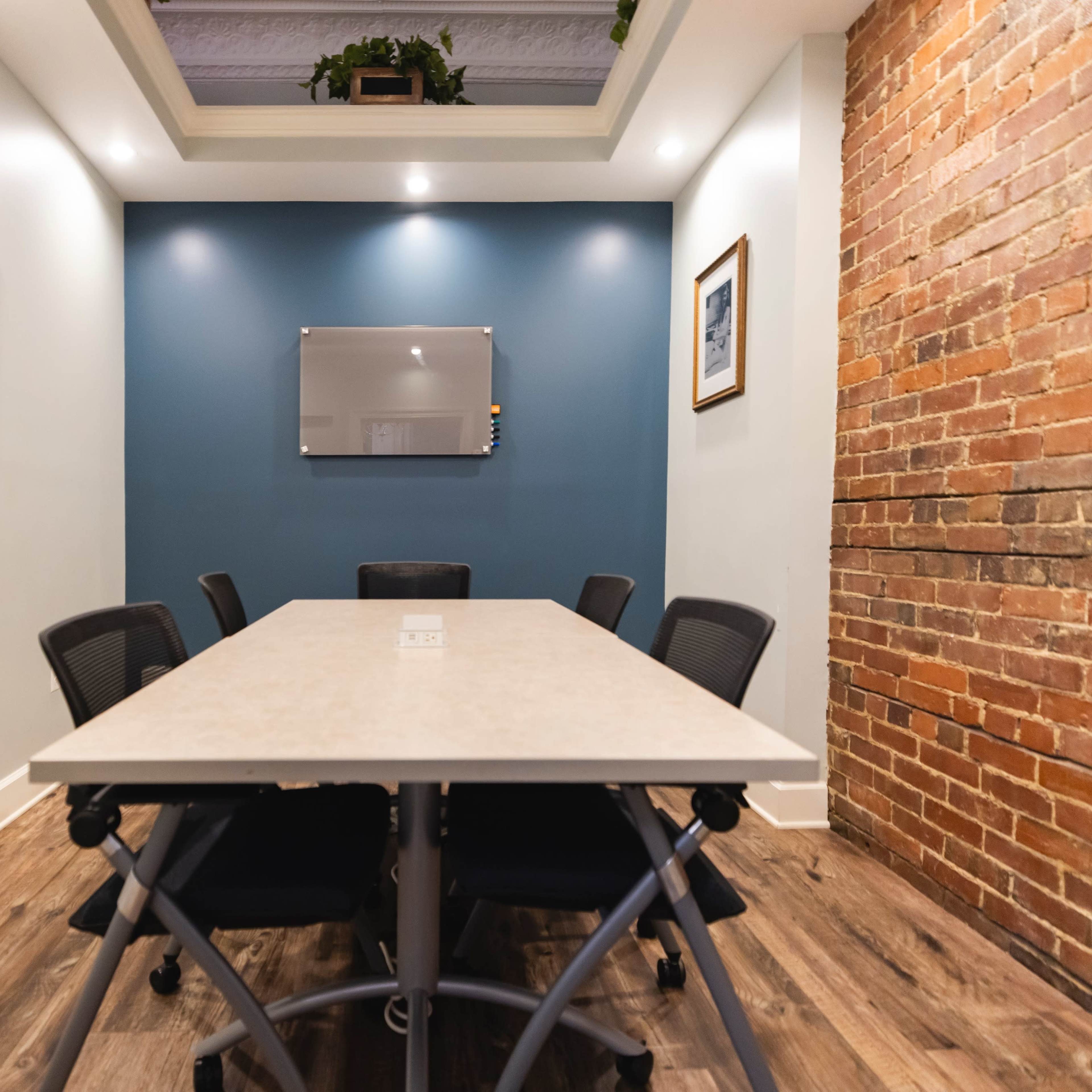 A meeting room features a long table surrounded by black chairs, with a wall-mounted screen and a blue accent wall.