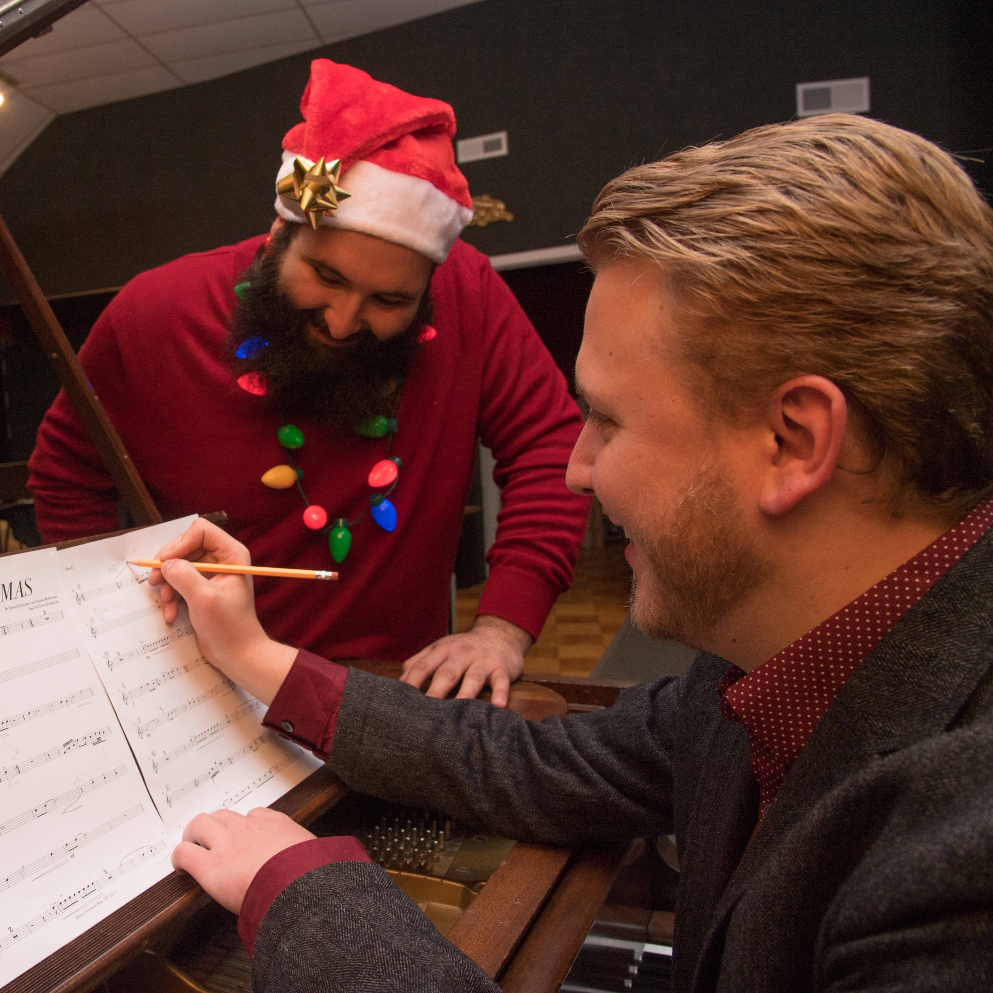 Two men are in a recording studio, with one wearing a Santa hat and festive sweater, as they look over sheet music on a grand piano.
