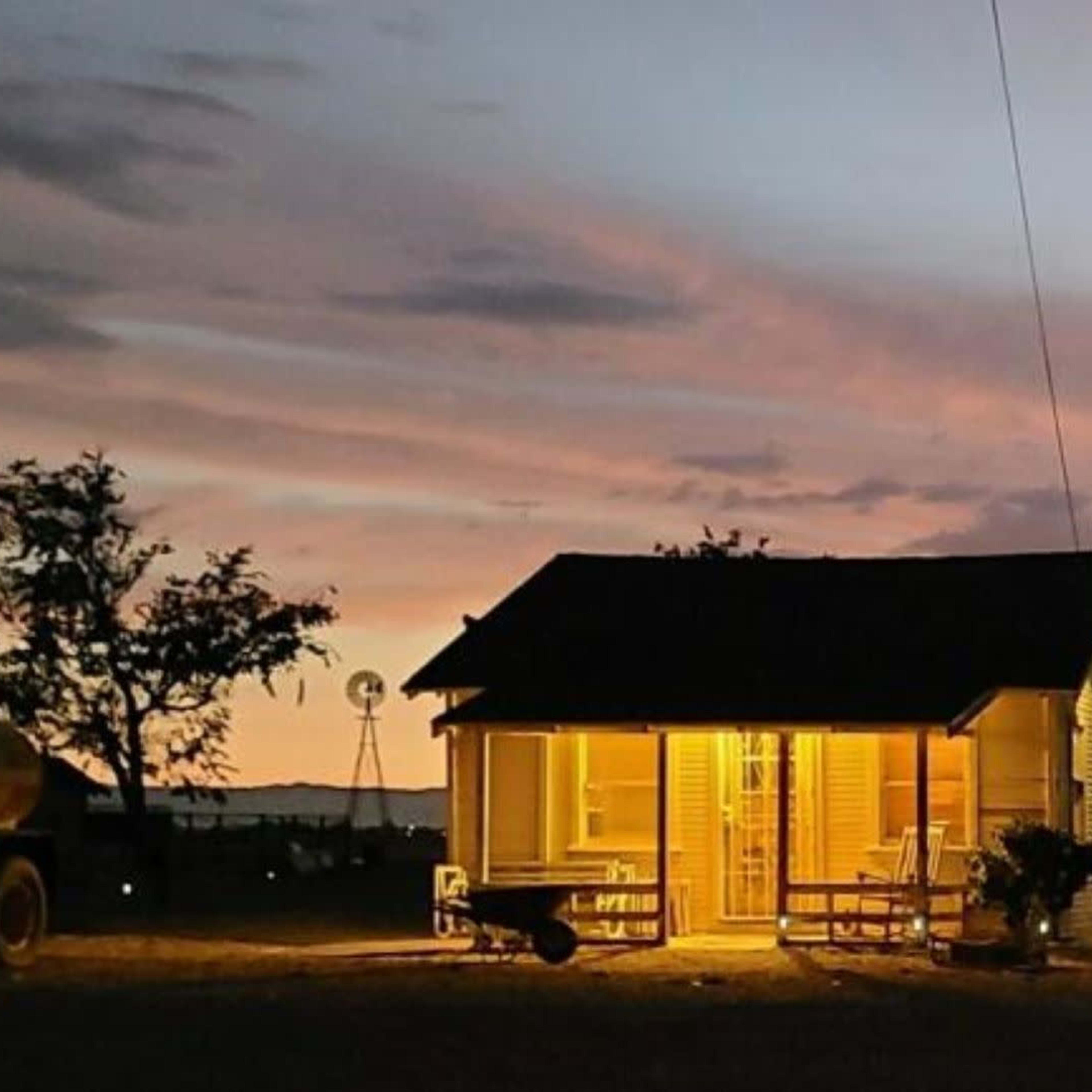 A small house with illuminated windows sits against a colorful sunset, surrounded by trees and a silo in the distance.