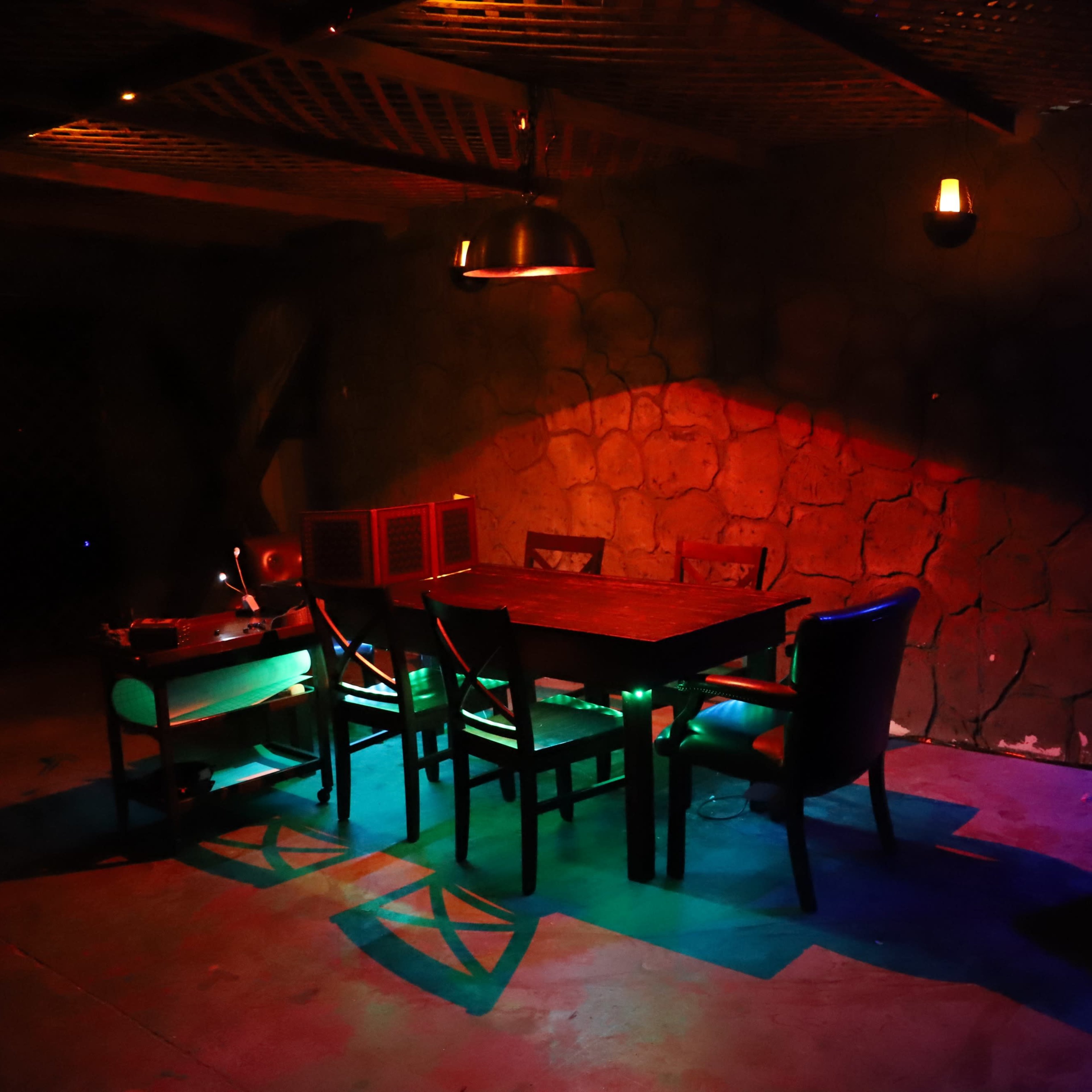 A dimly lit dining area features a large wooden table surrounded by chairs, with colorful lights casting shadows on the stone walls.