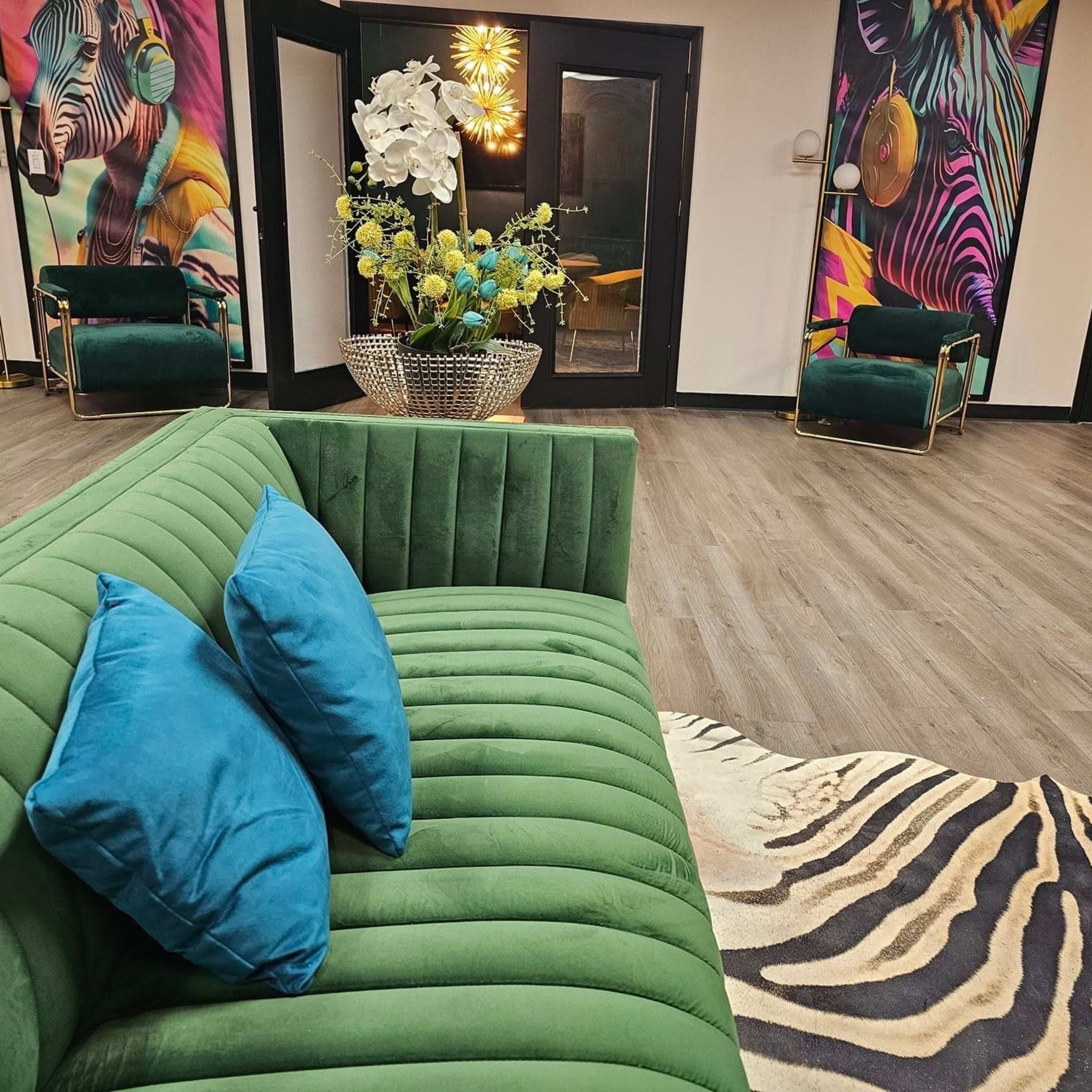A modern interior with a green velvet couch, two stylish chairs, vibrant wall art, and a floral arrangement in a decorative vase on a wooden floor.