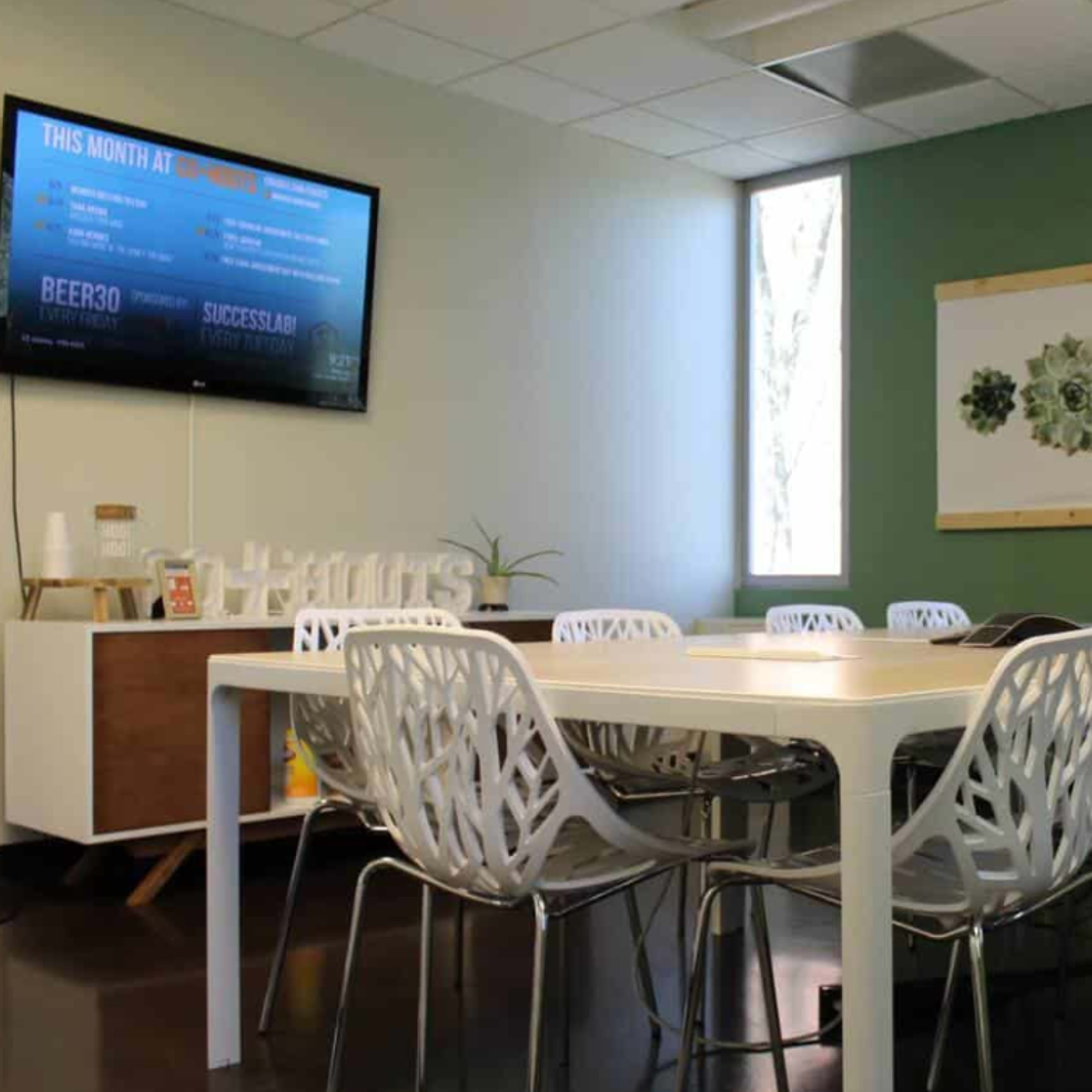 A modern meeting room with a large table surrounded by white chairs, a television mounted on the wall, and natural light coming through two windows.