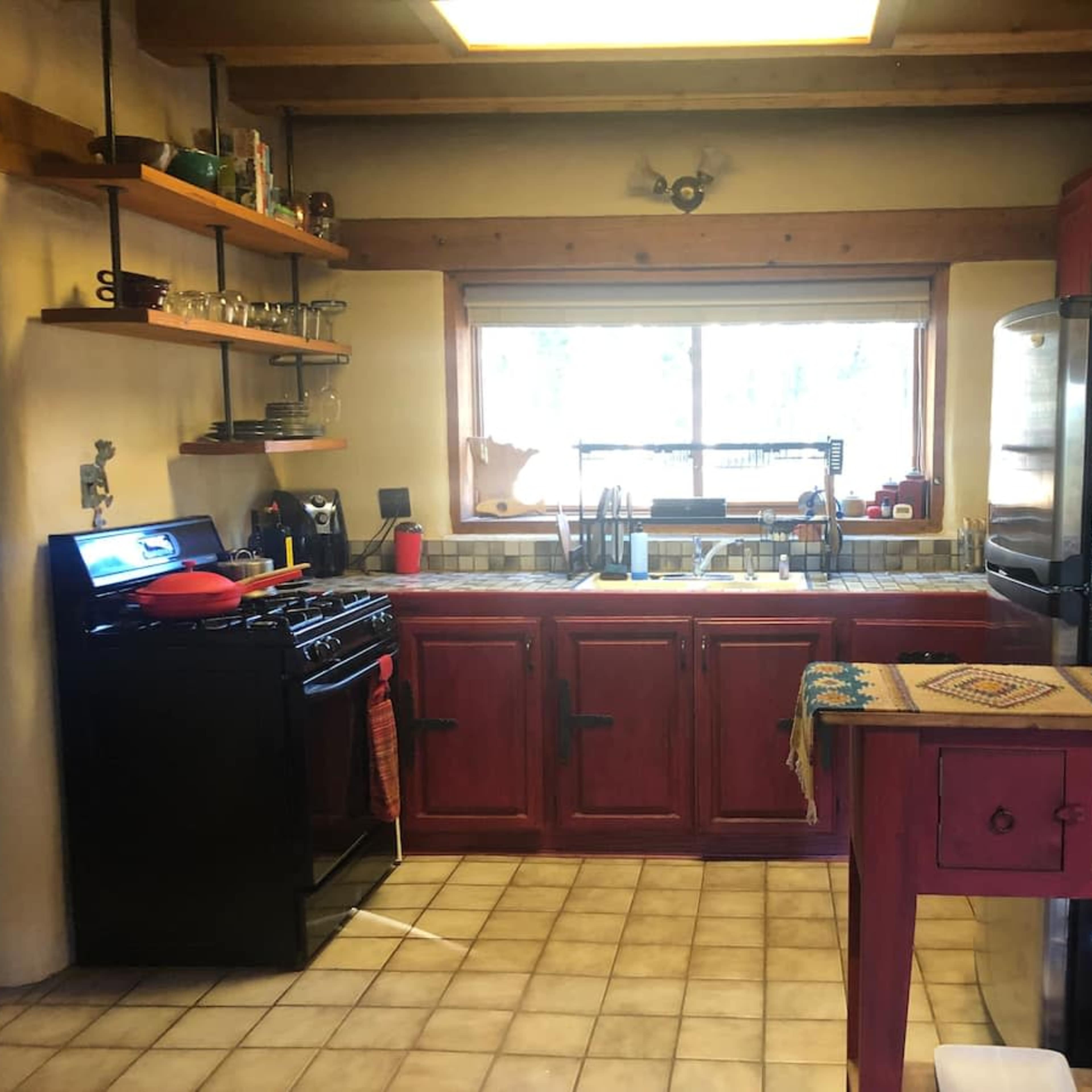 A rustic kitchen features red cabinetry, a gas stove, a large window, and a black refrigerator, with a table and shelves displaying various kitchen items.