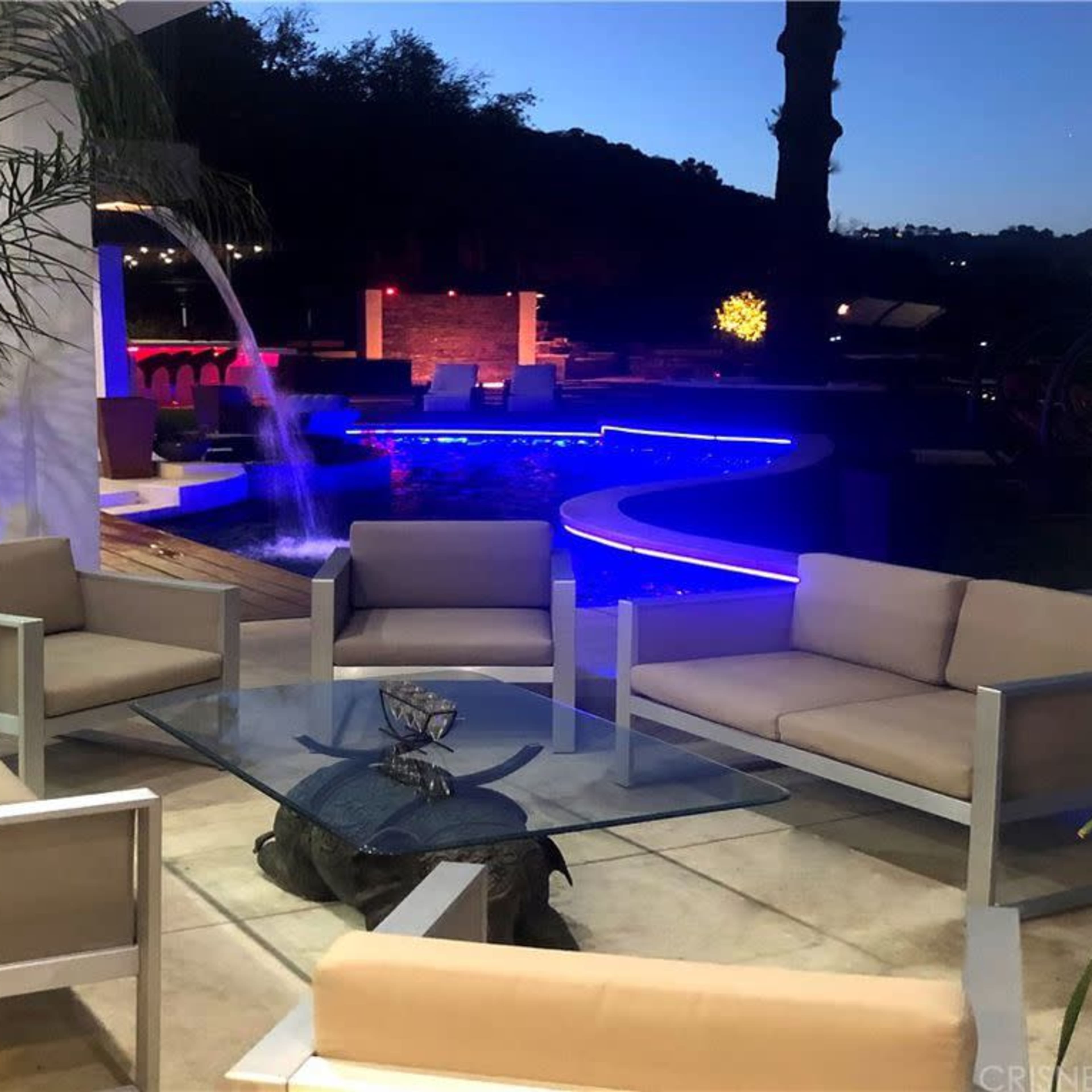 A modern outdoor seating area with beige sofas arranged around a glass table, next to a pool with colorful lighting and waterfalls.