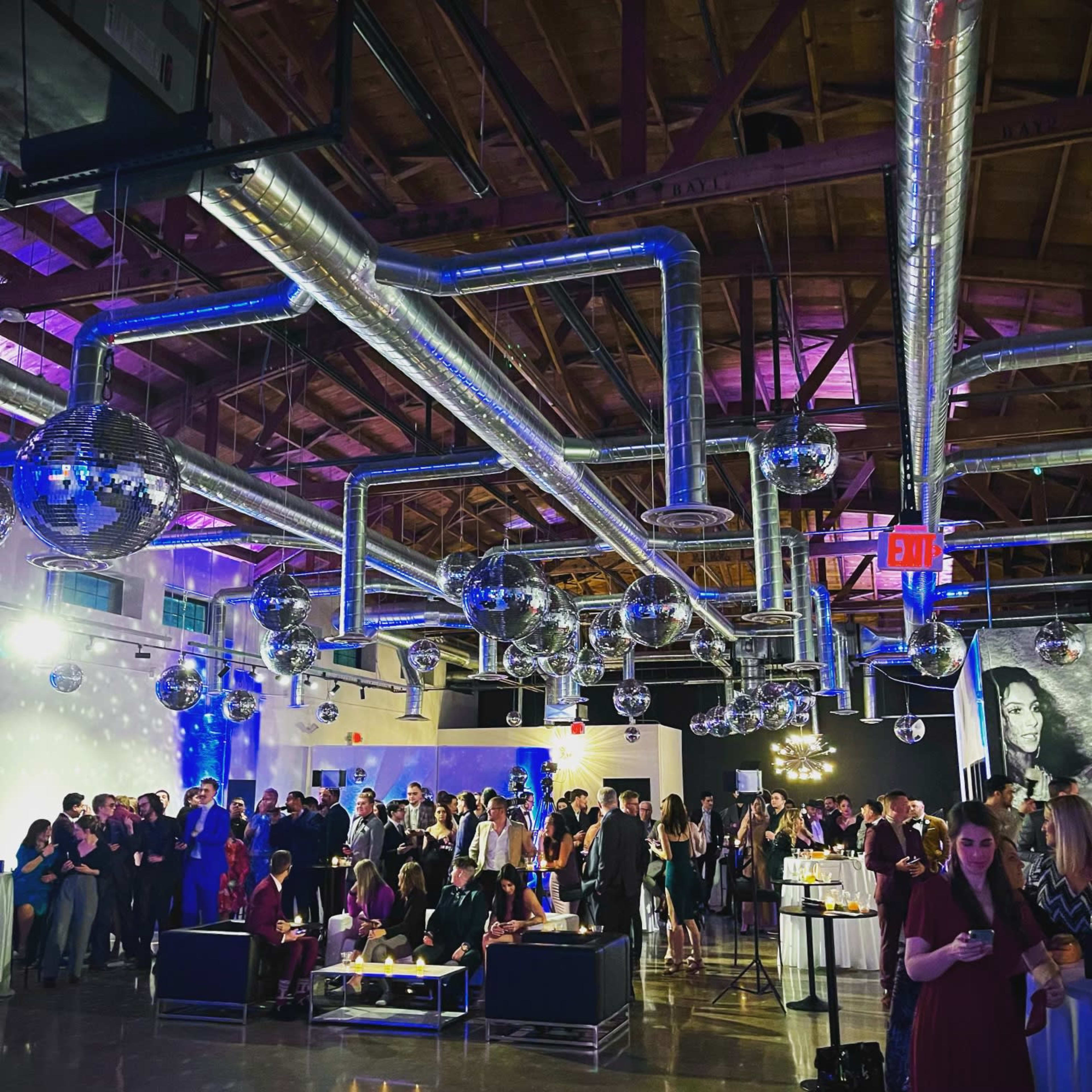 A large gathering takes place in a stylish venue featuring disco balls, exposed ductwork, and people mingling around tables.
