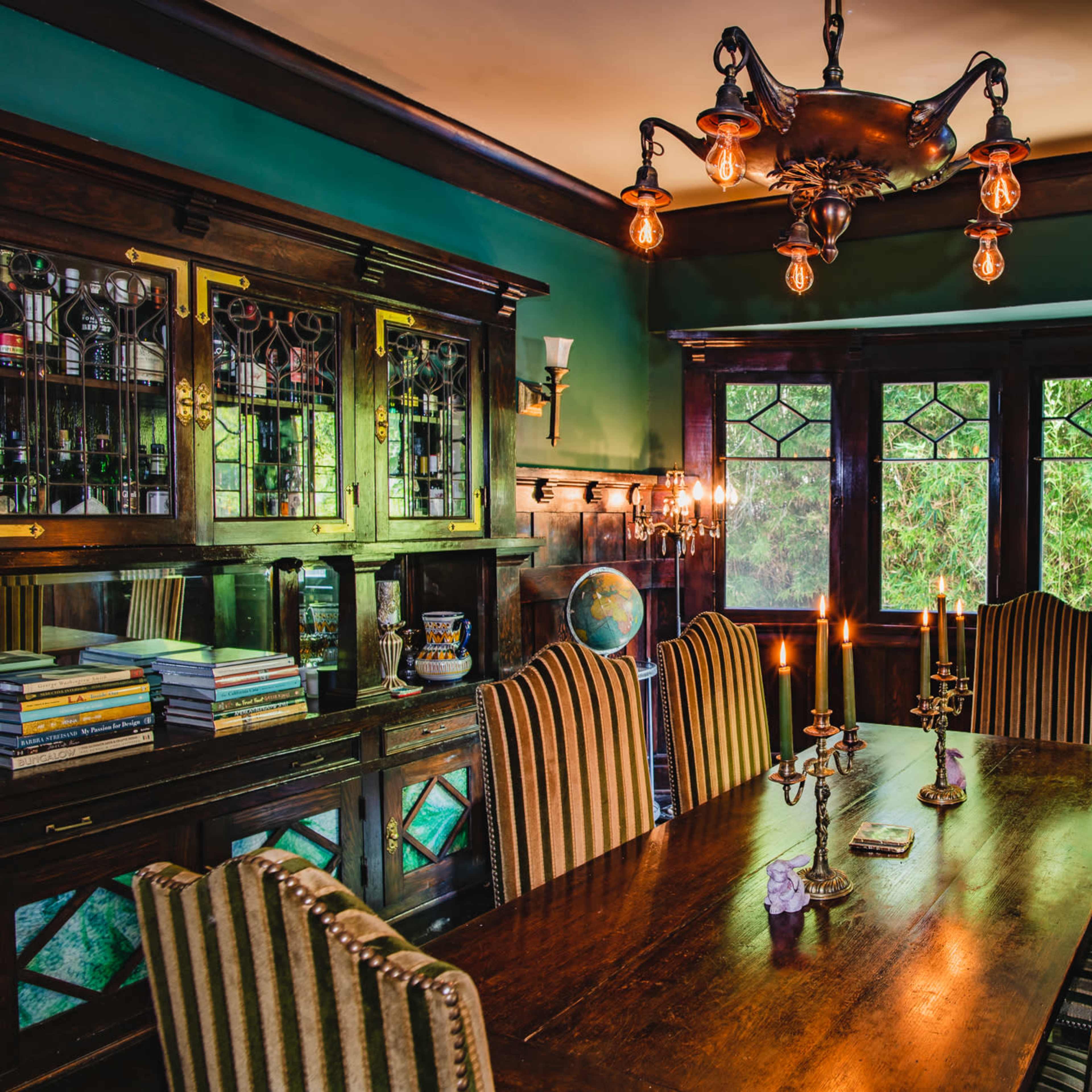 A dimly lit dining room with a long wooden table, striped chairs, and vintage lighting fixtures, accompanied by a cabinet displaying books and decorative items.