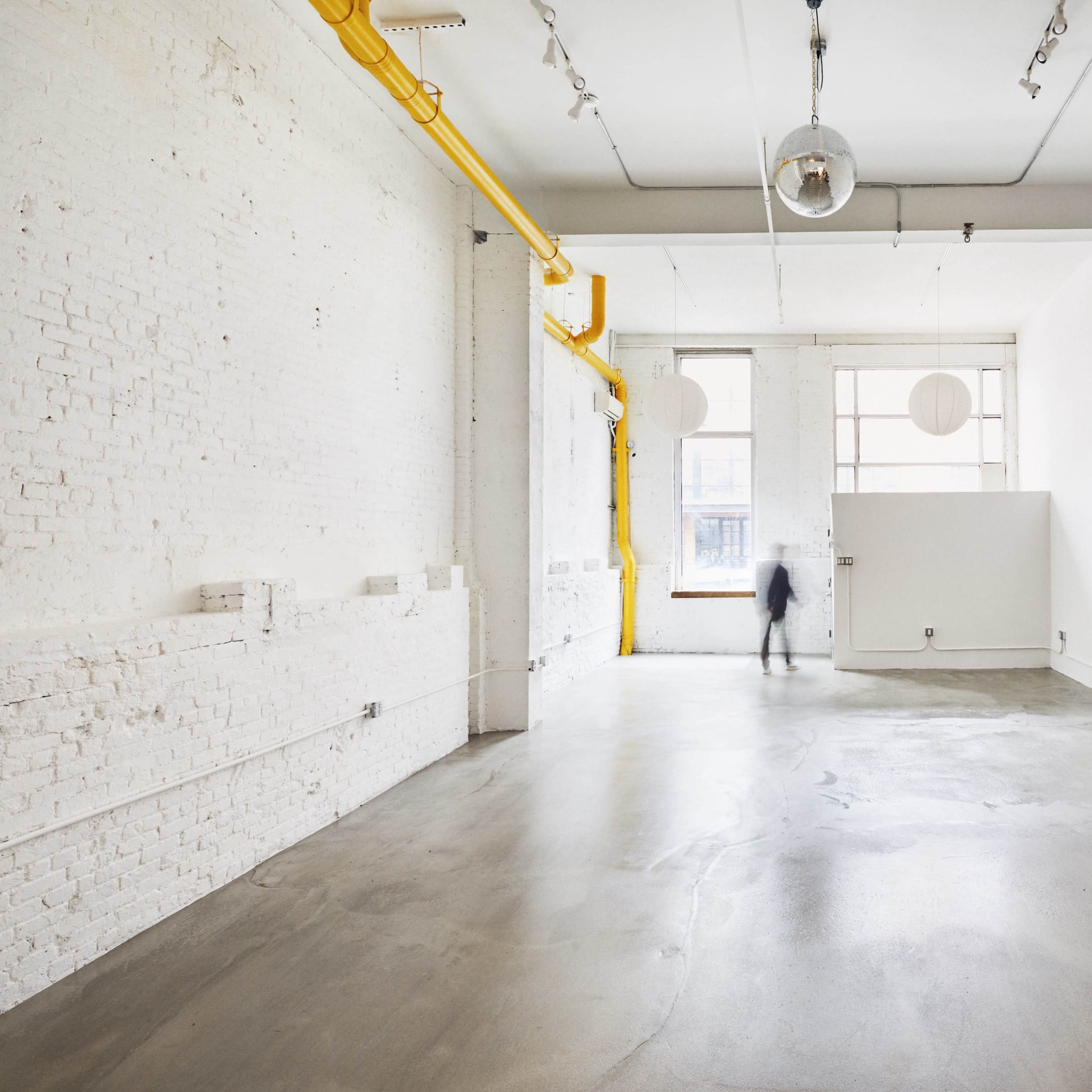 A spacious, empty room with white brick walls, a concrete floor, and large windows allowing natural light to enter.
