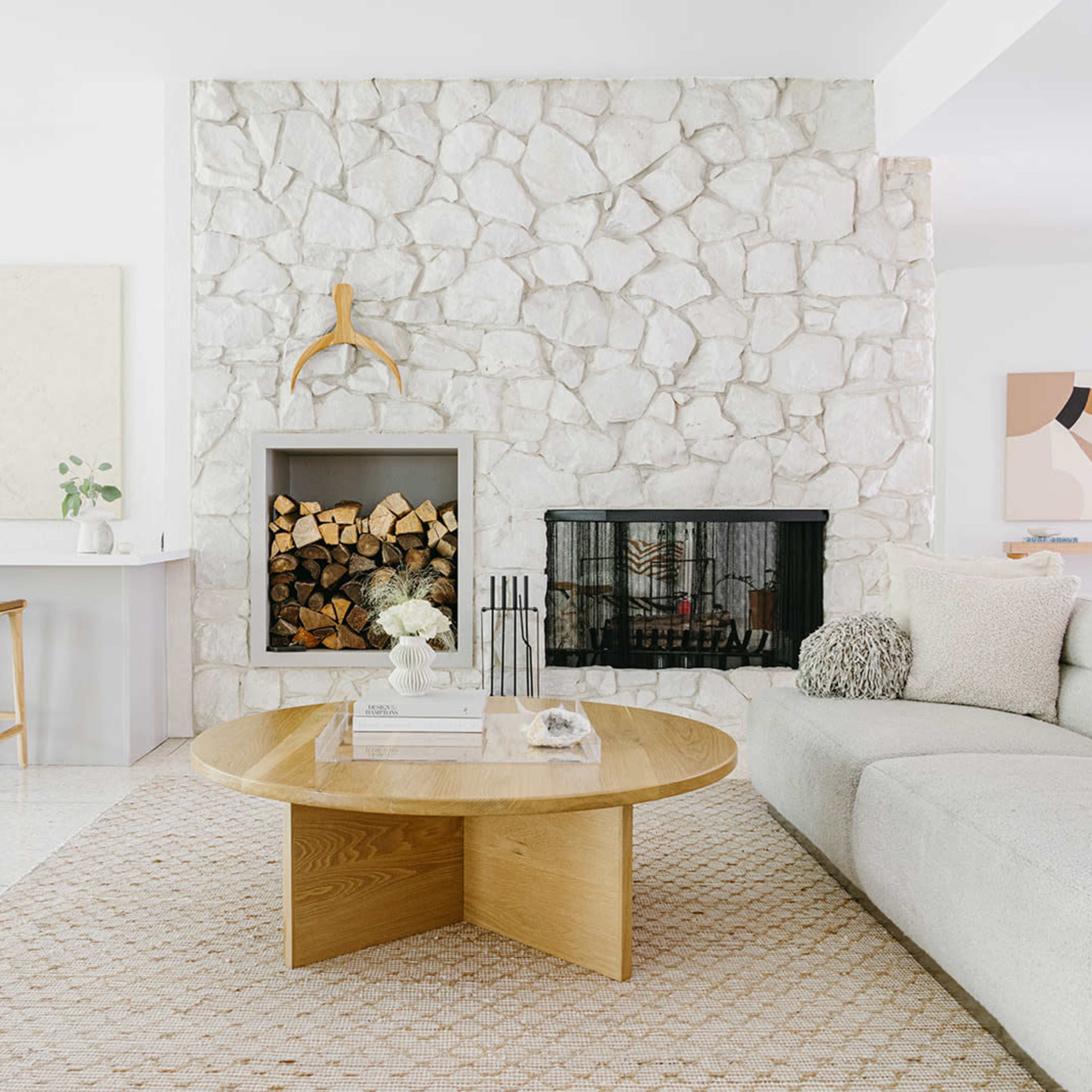 A light-filled living room featuring a stone fireplace with neatly stacked firewood, a beige circular coffee table, and a sectional sofa.