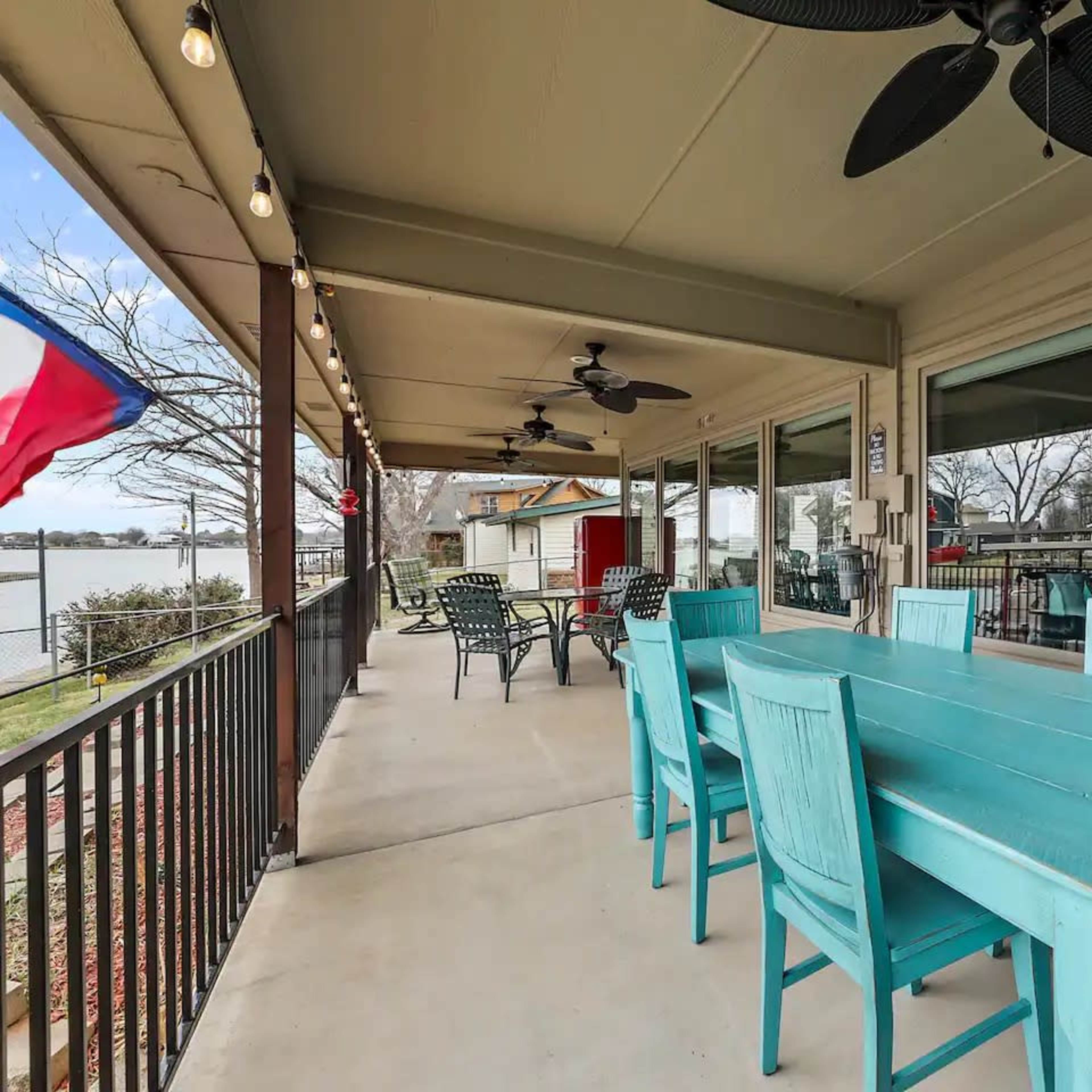 A covered patio features a large turquoise table surrounded by matching chairs, with a view of a lake and nearby homes.