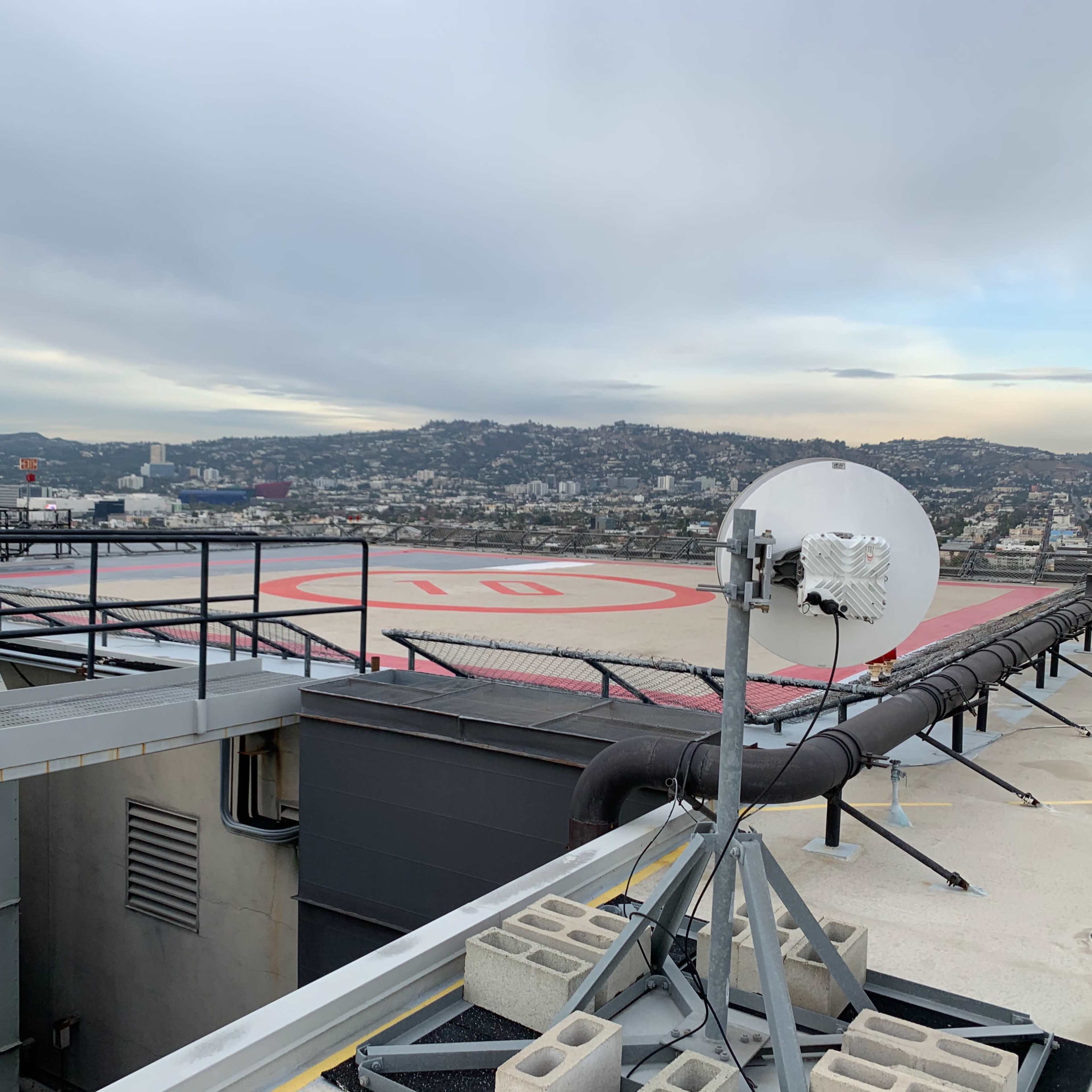 A rooftop helipad overlooks a cityscape with hills in the background and a satellite dish mounted nearby.