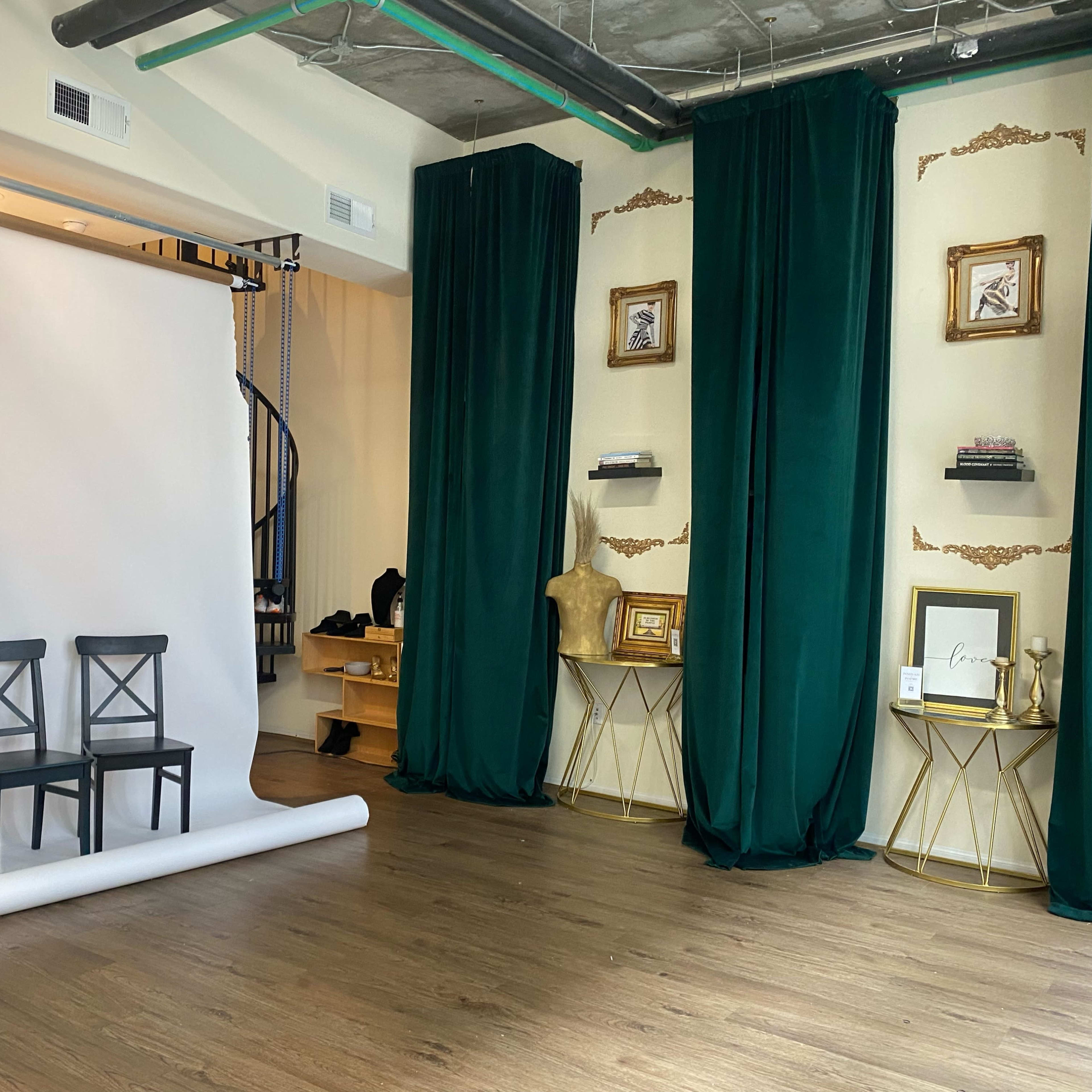 A photography studio featuring three black chairs in front of a white backdrop, flanked by green curtains and gold-framed decorations on the walls.