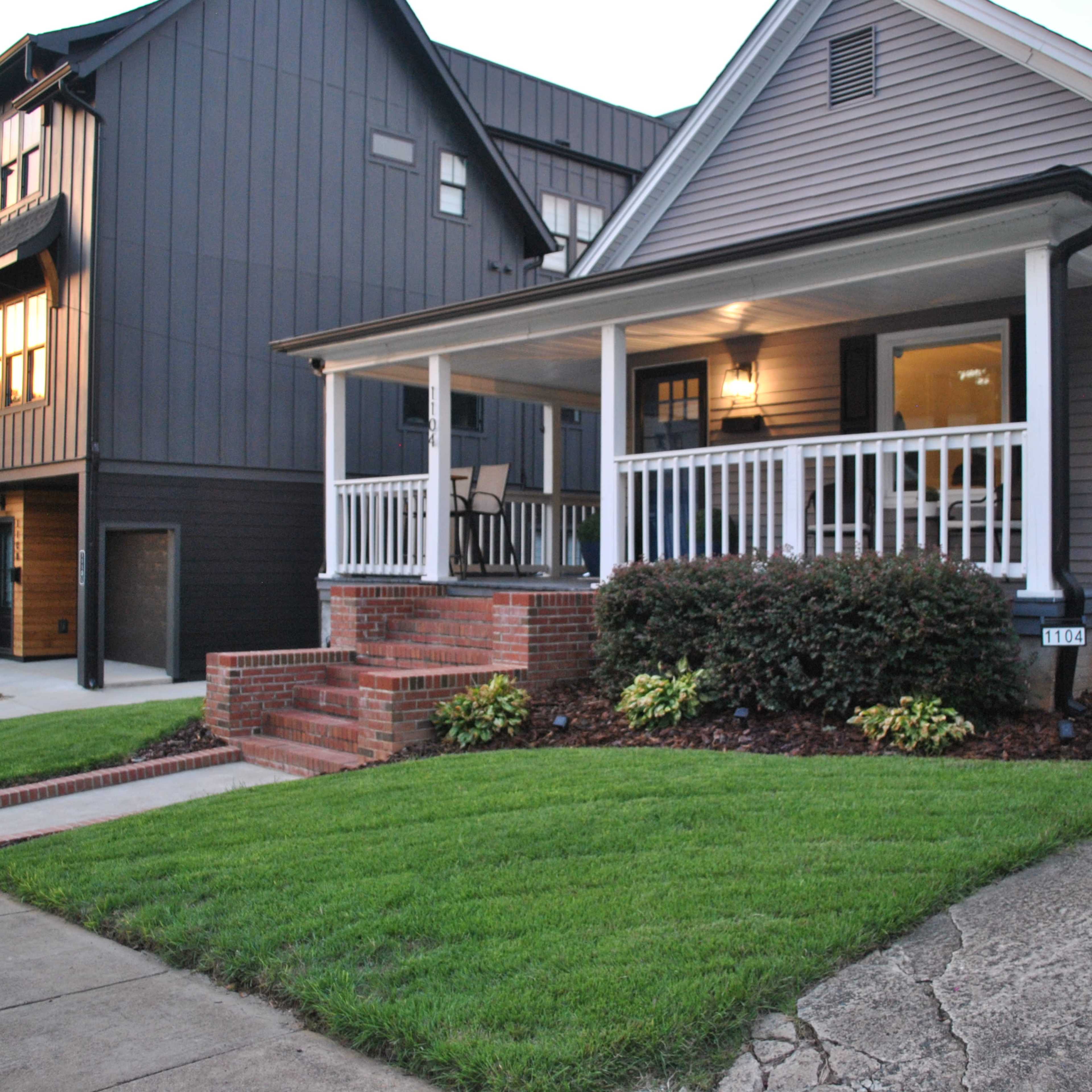 A residential area featuring a gray home with a porch and brick steps, alongside a modern dark-colored house, with well-maintained lawns and sidewalks.